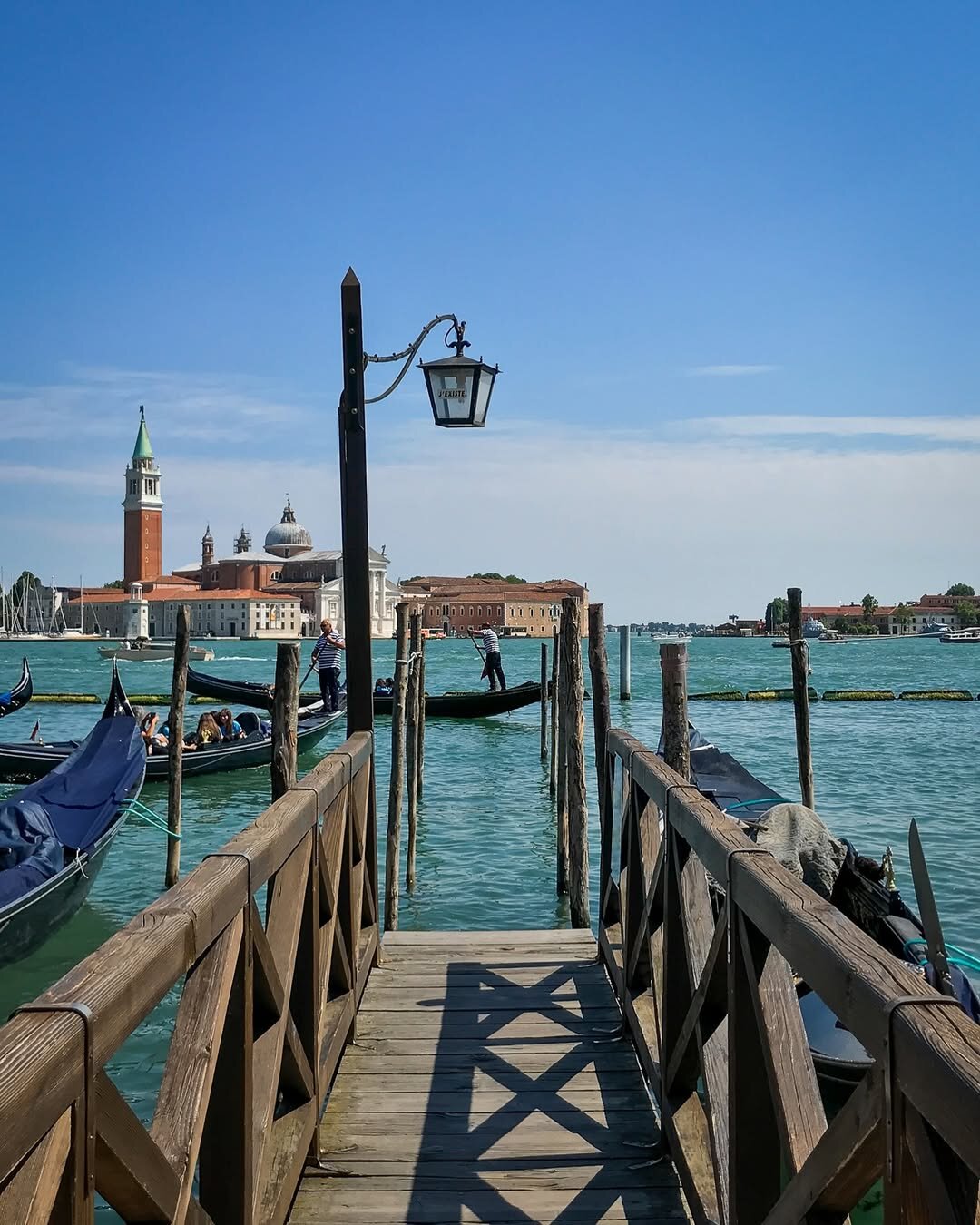 Wooden dock in Venice with gondolas moored on both sides, vintage lantern labeled “Venezia,” and the Church of San Giorgio Maggiore with its bell tower in the background under a clear blue sky. #venice #italy #canals #gondola #sangiorgiomaggiore #lagoon #travel #photography #fotografia #architecture #renaissance #watercity
