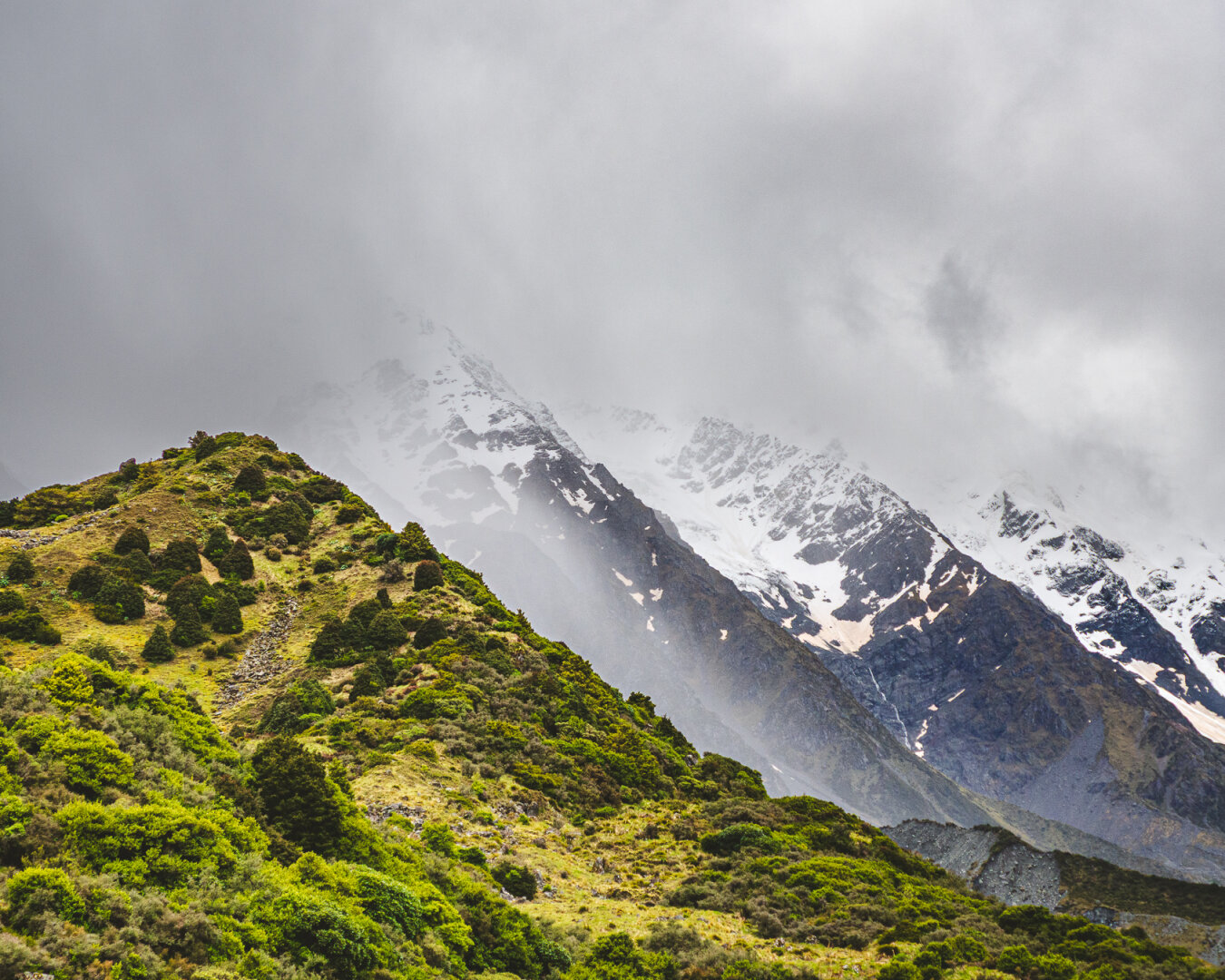 A green, shrub-covered mountain ridge rises in the foreground, with steep snow-covered peaks behind it. Heavy gray clouds hang low over the mountains, partly obscuring the highest summit.