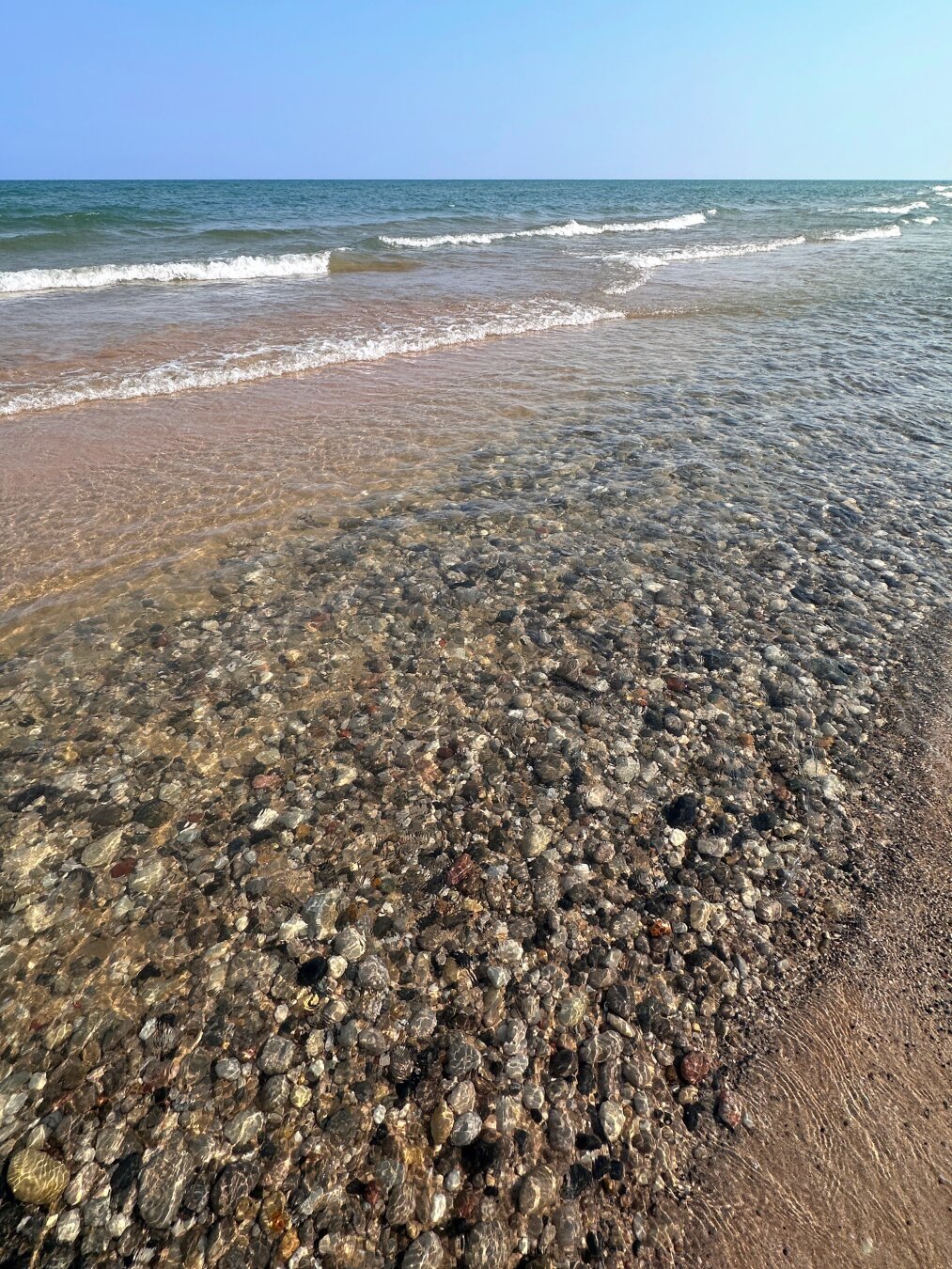 A picture of the Lake Michigan shore with clear skies and waves. A large swath of rocks lie just beneath the waters surface in a channel along the beach.