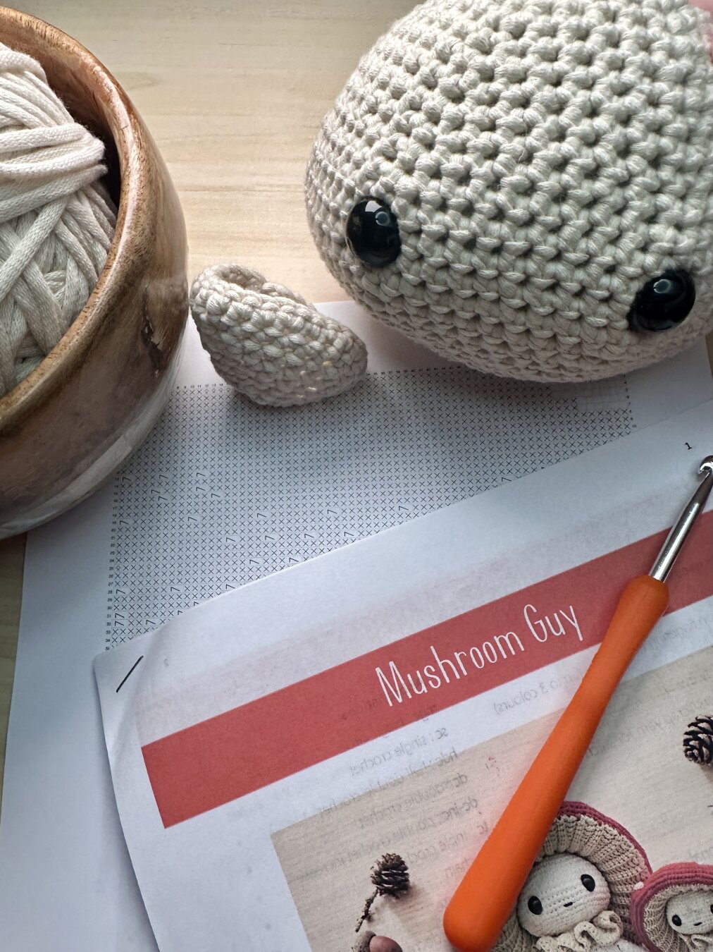 Photo of a crochet project in progress. Using Bernat Home Dec yarn to create a Mushroom Guy plushie. Includes stuffed head piece with two black eyes, one unstuffed leg, a ball of yarn in a wooden bowl, crochet diagrams, instructions, and an orange crochet hook.