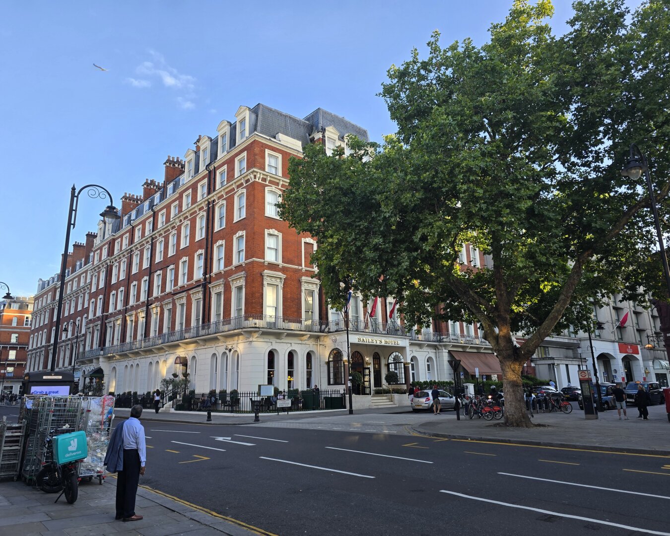 A quiet street in the foreground ironically contrasts with the crowded market behind the photographer (not pictured). Across the street there's a large tree full of green leaves in the middle ground, and a four story building of red brick in the background.