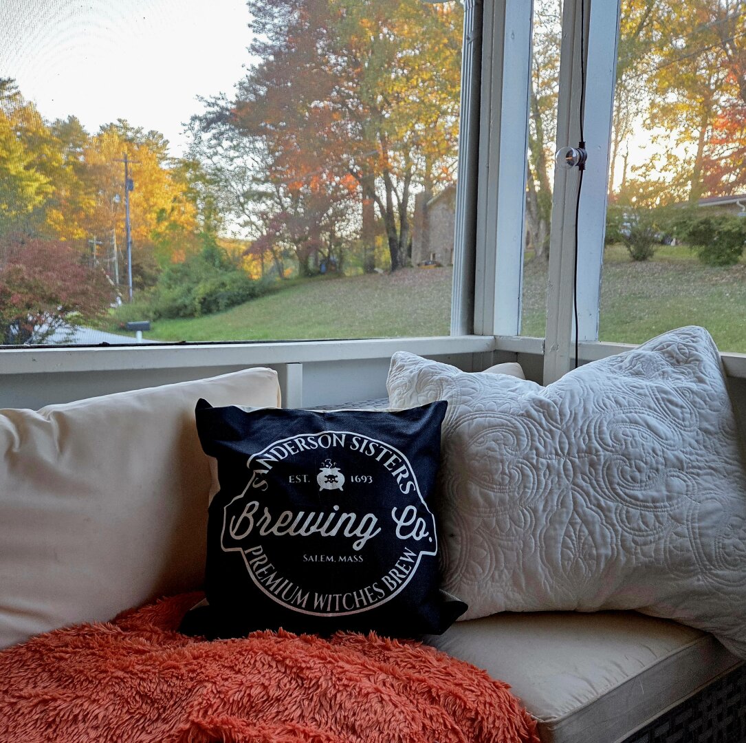 A cozy screened porch in the Appalachian Mountains during peak October color. A cushioned bench holds a white quilted pillow, an orange fuzzy throw, and a black pillow that reads “Sanderson Sisters Brewing Co. – Premium Witches Brew – Salem, Mass.” Outside the porch, the hills and trees glow in warm shades of gold, orange, and red under soft evening light. The scene feels peaceful and autumnal, evoking the comfort of mountain living in fall.