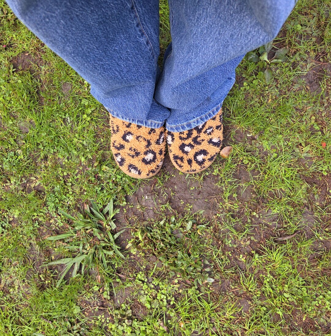 You’re looking straight down at a pair of feet standing on a patchy grassy yard. The person is wearing blue jeans that cover most of their legs and slightly bunch around the ankles. On their feet are fuzzy, warm-looking slip-on shoes with a bold orange-and-black leopard print pattern. The ground beneath them is a mix of bright green grass, small weeds, and patches of bare brown soil, giving a slightly damp, uneven, natural feel — like a backyard or garden area that’s a little muddy in spots.
