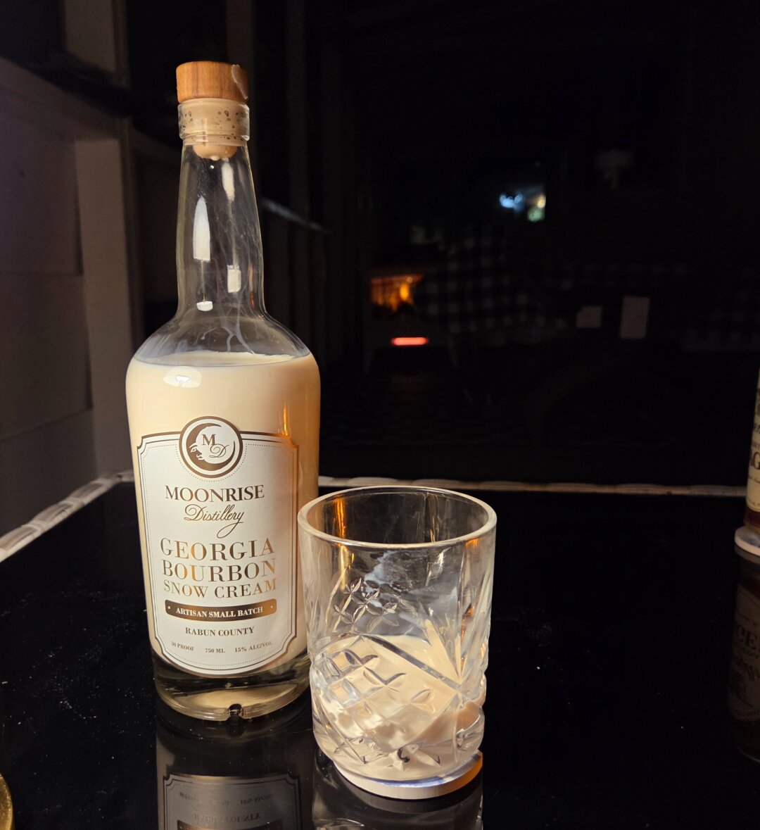 The image shows a cozy nighttime porch scene. On a glossy dark tabletop sits a bottle labeled “Moonrise Distillery, Georgia Bourbon Snow Cream”, produced in Rabun County. The bottle has a pale wooden cork and contains a creamy, light beige liqueur. Next to it is a short, faceted glass with a small amount of the same creamy drink inside, catching warm reflections from nearby light. The background is dimly lit, with hints of a checkered tablecloth and a soft amber glow, evoking a calm Appalachian evening perfect for sipping something sweet and strong.