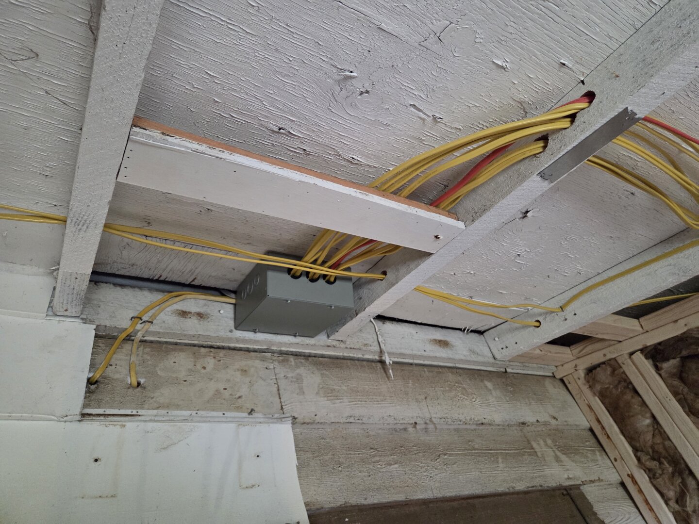You’re looking up at the unfinished ceiling of an old house where electrical work is in progress. The ceiling is made of wide white-painted boards and exposed wooden joists. Several bright yellow electrical cables (and a couple of red ones) are neatly threaded through holes drilled in the joists, running across the ceiling like organized veins.

In the center-left of the image, there’s a gray metal electrical junction box mounted to a beam, with multiple yellow wires feeding into it. The surrounding area shows a mix of old wood and newer framing .  The wall below still has rough, weathered boards with faded white paint, while to the right you can see new pale wood studs and some brown insulation tucked between them.

The overall scene feels like a mid-renovation moment with new wiring being carefully installed while much of the old structur