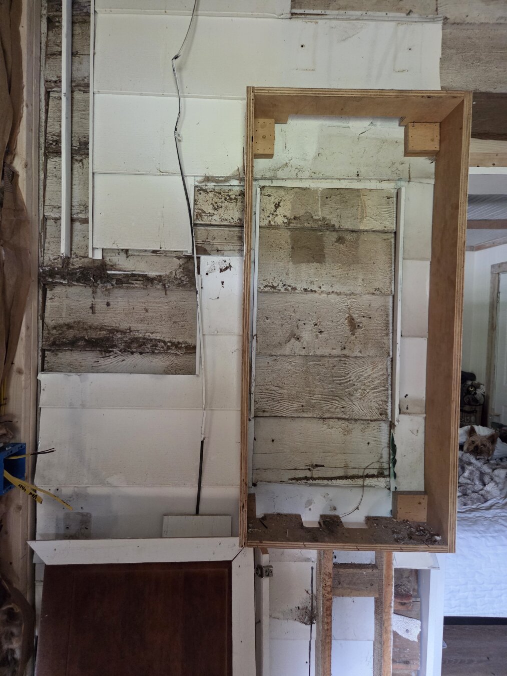 You’re looking at an unfinished interior wall of an old house where renovation work is happening. The wall is built of white-painted 100+ year old wood, but the paint is worn and stained in places, and some sections have been removed or cut away, revealing rough, weathered wooden boards underneath.

Near the middle-right side of the image, there’s a rectangular wooden frame fastened to the wall.  It is the shell of a cabinet that needs removed. Inside the frame you can see the exposed old wood planks of the original wall. The bottom of this framed box is open with two vertical wooden studs or braces sticking down.

To the left, there’s a loose white electrical wire that snakes down the wall and hangs slightly. A blue electrical box with yellow sheathing peeks out on the far left edge. Below, there’s a detached cabinet door or panel leaning against the wall.

In the far background through an open doorway, you can see part of another room: a cozy bedroom with white bedding, with a Yorkie