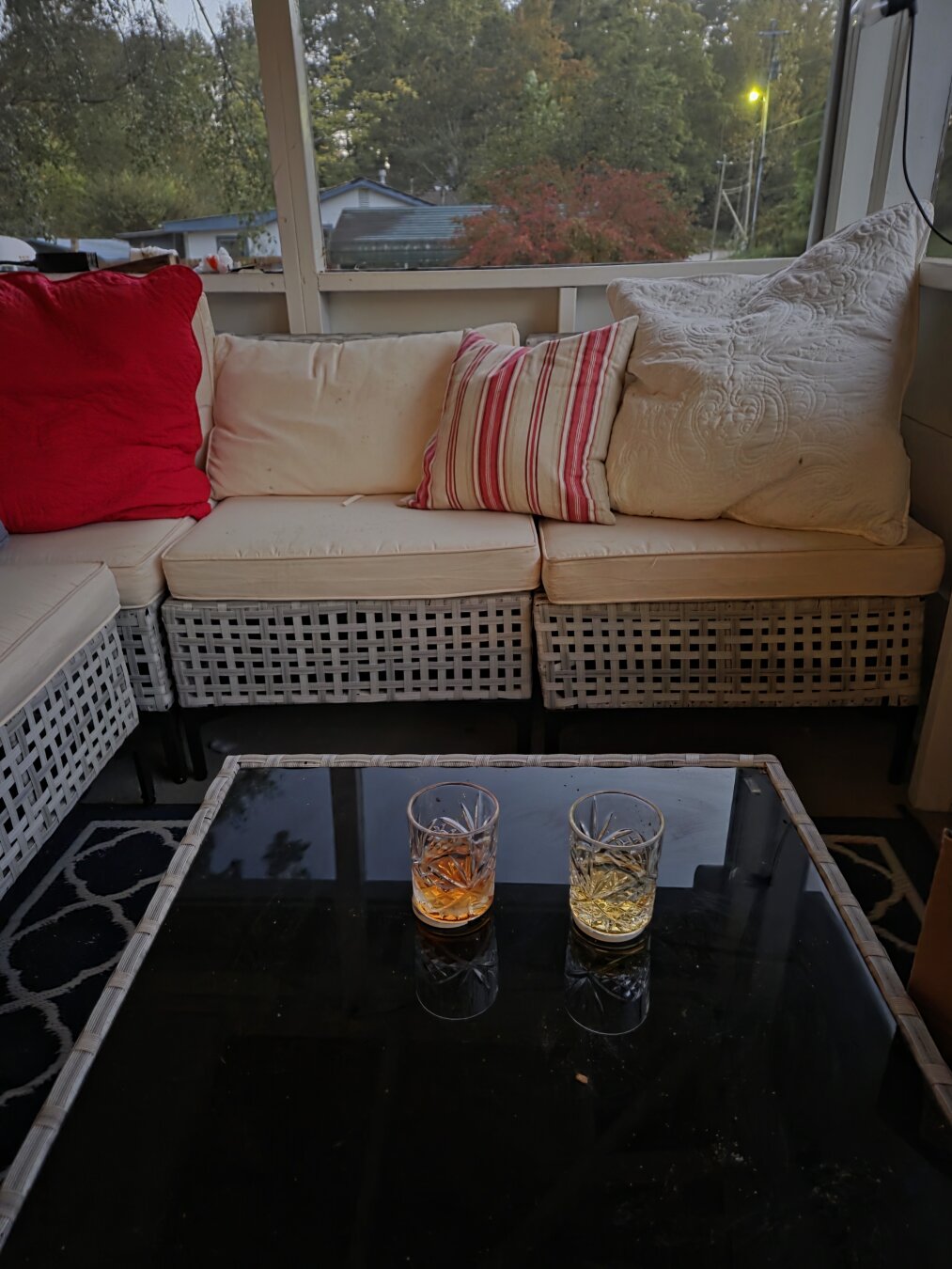 The photo shows a cozy screened-in porch with a relaxed, inviting feel. At the center is a square wicker coffee table with a shiny black glass top. On it sit two short, cut-glass tumblers — one filled with a deep amber drink and the other with a lighter golden drink, both catching the evening light and reflecting on the table’s surface.

Behind the table is an L-shaped wicker sectional couch with cream cushions and a mix of pillows: a big red pillow, a soft beige one, a red-and-white striped pillow, and another light pillow with a quilted texture. The couch sits on a navy blue rug with a white trellis pattern. Outside the screened porch, you can see autumn trees — one turning a rusty red, others still green — and a faintly glowing streetlight as dusk sets in. The whole scene feels warm, rustic, and perfect for sipping drinks on a cool Appalachian evening.