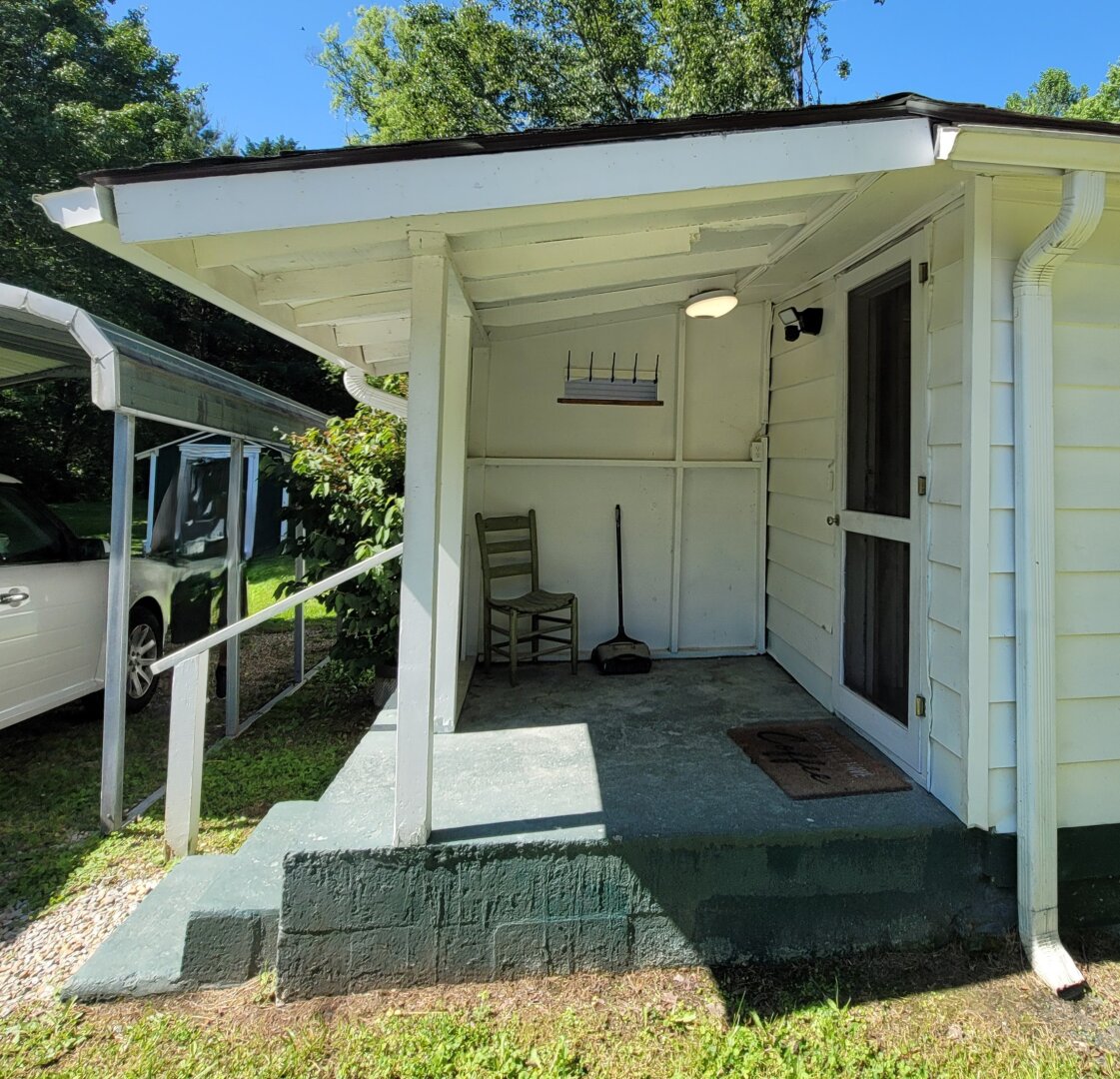 The photo shows the “before” version — an open back porch attached to a small white house. The roof is supported by posts, and the porch has a simple concrete floor with steps leading down to the yard. A wooden chair, broom, and dustpan sit along the back wall. To the right is a screen door leading into the house. The porch looks exposed to the weather and minimally used.
