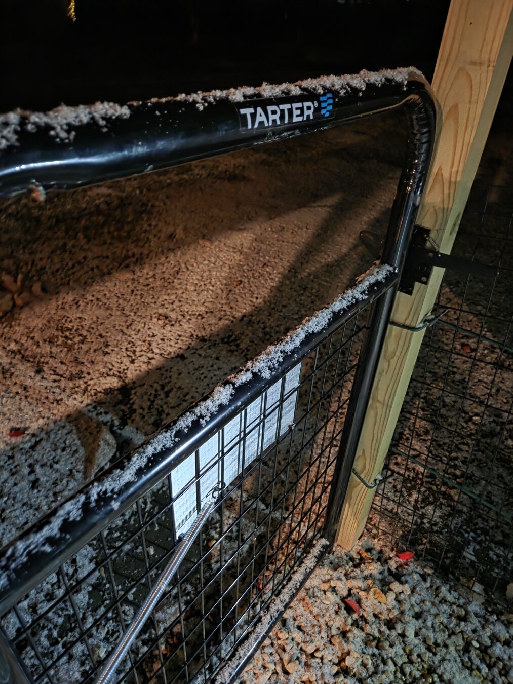 Snow on a Gate
This is a nighttime photograph showing a black metal utility gate dusted with a light layer of snow or sleet.
 * The Gate: The gate is made of a black, rounded, tubular metal frame with a square metal mesh/wire infill. The upper horizontal rail of the gate clearly displays the manufacturer's name, "TARTER®," in white lettering.
 * The Snow: A thin, white, granular layer of snow rests on the top surface of the gate's frame and is also scattered thickly on the ground visible through the gate.
 * The Setting/Ground:  The ground is covered with what appears to be light-colored gravel or crushed stone, which is heavily mixed with the same white snow/sleet. The overall impression is a mix of brown/tan earth and white snow.
 * The Post: The gate is mounted to a tall, light-colored wooden post ( pressure-treated pine).