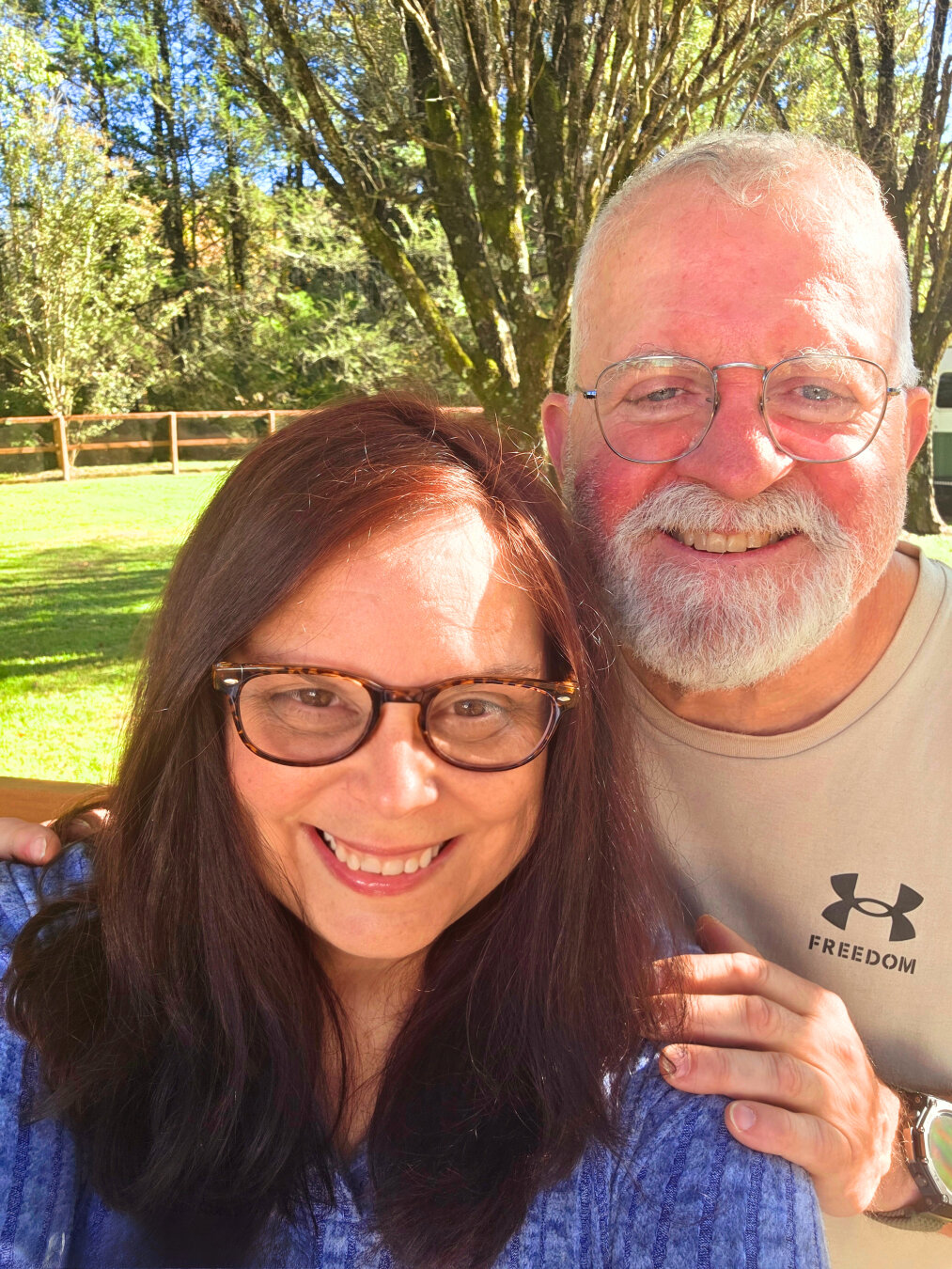 A smiling couple is standing closely together outdoors on a bright, sunny day in what looks like a peaceful rural or mountain setting. They are both wearing glasses and look happy and relaxed. The woman is in the foreground, with long dark red hair and a blue textured sweater, wearing no makeup. She has a warm smile and is holding the camera slightly above eye level for a selfie. The slightly sunburnt man stands just behind her with his hand resting gently on her shoulder. He has short white hair, a full white beard, and is wearing a beige T-shirt with a small Under Armour logo and the word “FREEDOM” on it. Sunlight highlights both of their faces, giving the image a cheerful, golden tone.

Behind them is a grassy yard bordered by a simple wooden fence and tall trees. The trees are full of leaves, suggesting early autumn with sunlight filtering through the branches. The scene feels calm, natural, and joyful.  A moment of contentment shared in a beautiful outdoor setting.