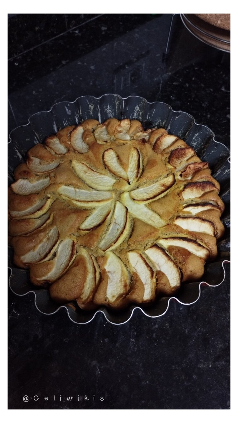 Tarta de manzana recién sacada del horno. Aún está en el molde cuyos bordes son ondulados. La tarta está dorada y tiene lascas de manzana dispuestos en forma de circunferencia (cerca del borde) y estrella (centro).