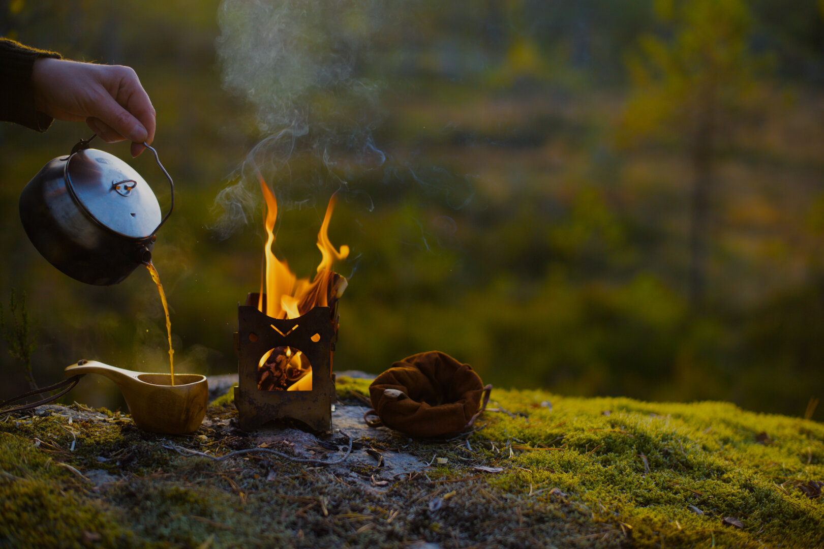 coffee is poured into a wooden cup from a kettle besides a burning firebox