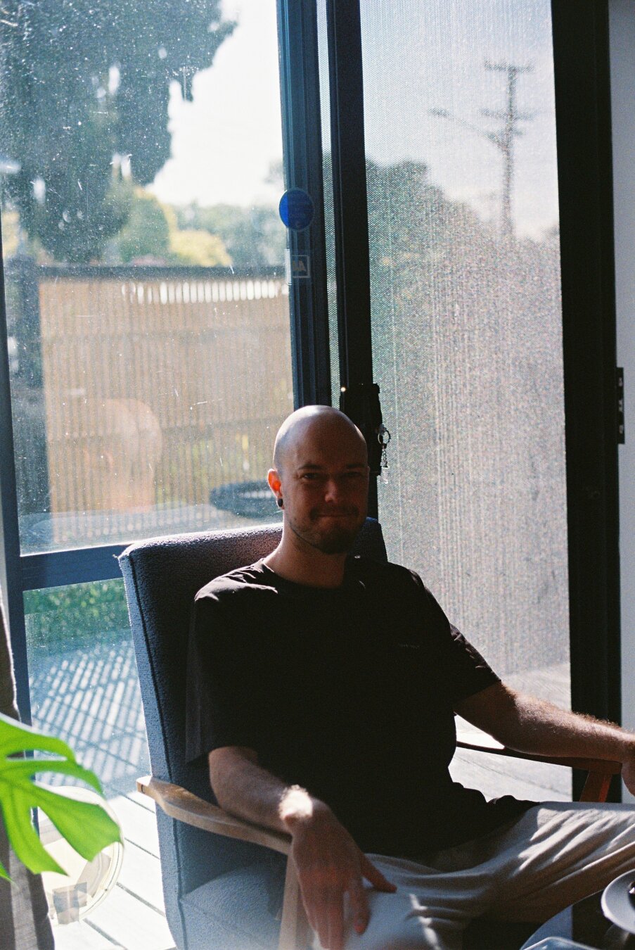 A bald dude sitting in an old chair in front of a glass door looking at the camera.