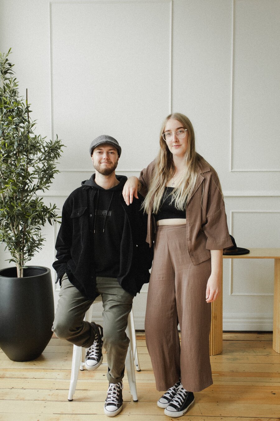 A couple in a studio with a wooden floor, white walls, and a tall plant, with the man sitting on a stool.