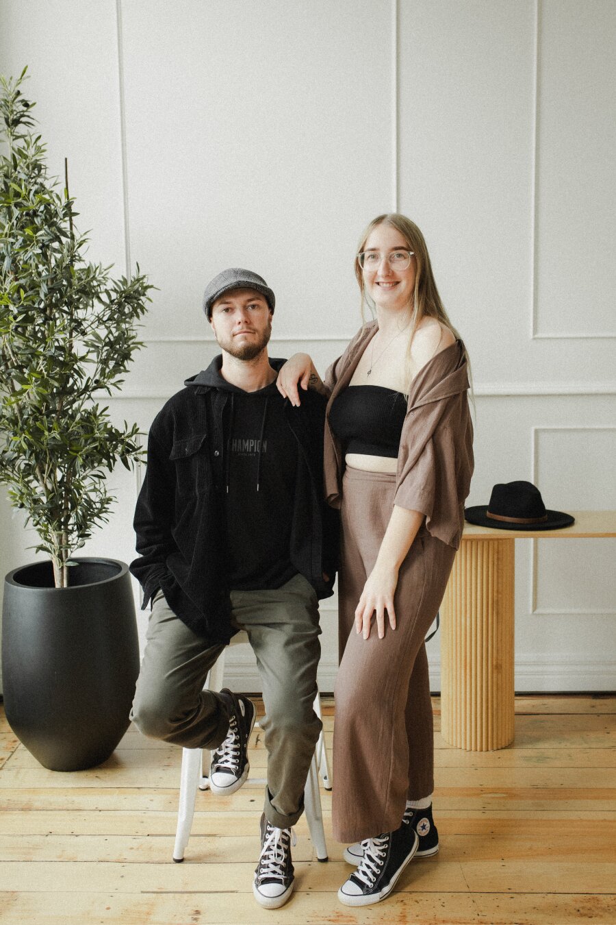 A couple in a studio with a wooden floor, white walls, and a tall plant, with the man sitting on a stool.
