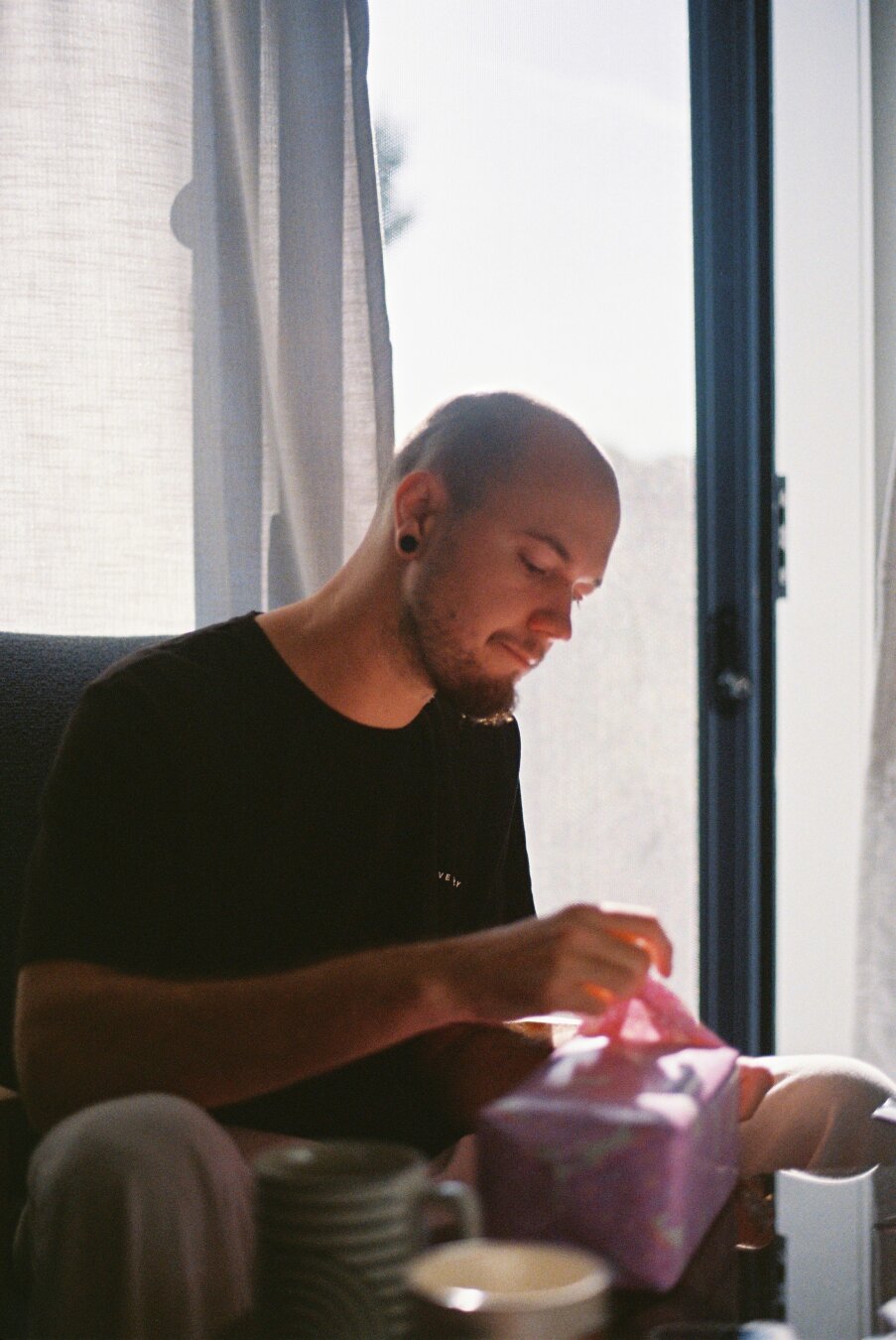 A bald dude sitting and opening a gift.