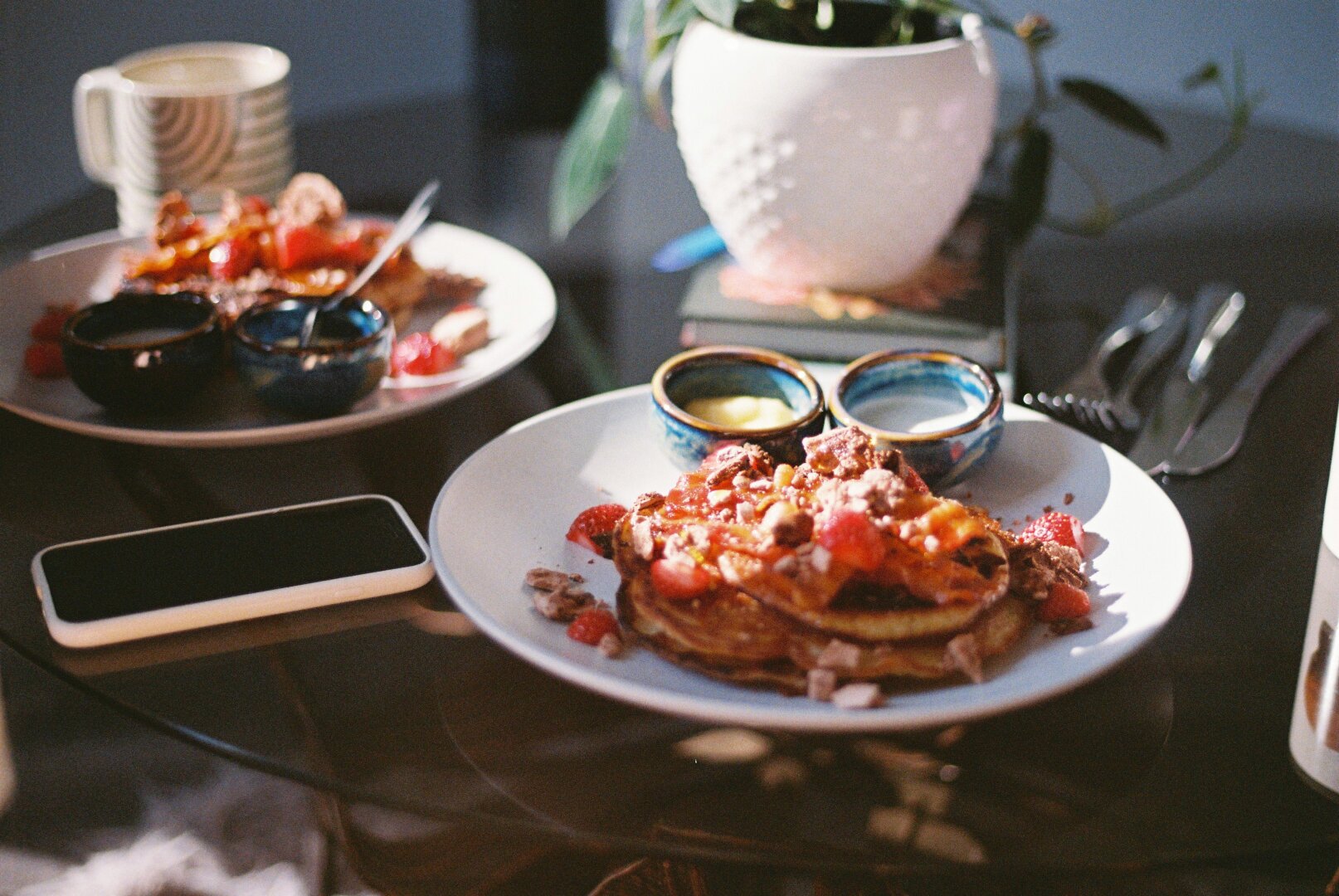 Two plates with pancakes loaded with berries, and ramekins, a phone, and a mug on a glass table.