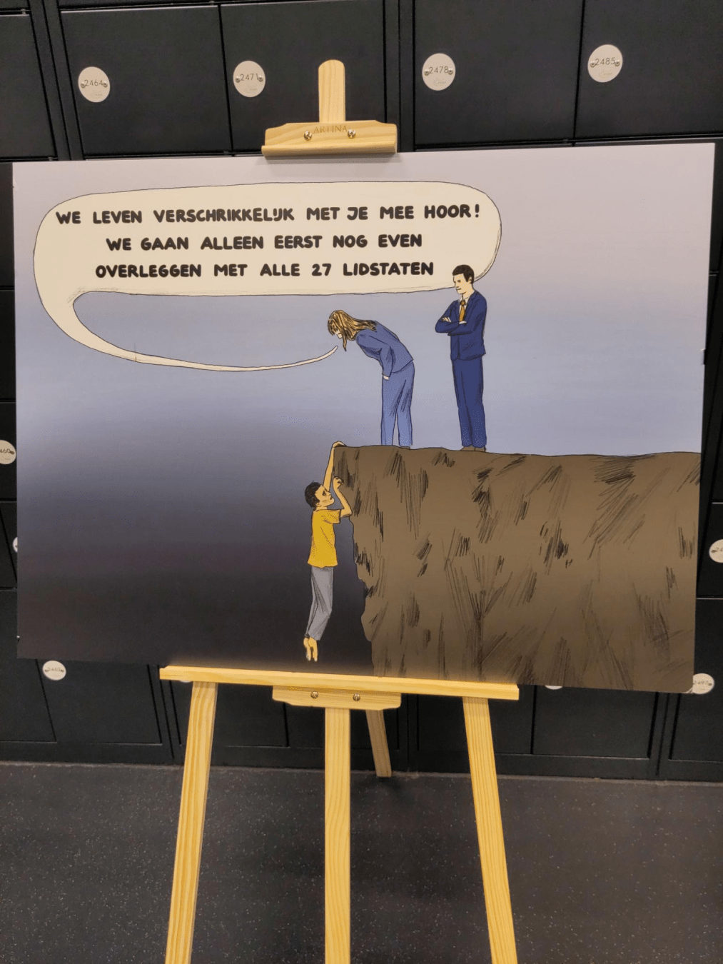 Geschilderde comic op een schildersezel. Een persoon hangt aan een klif. Twee mensen staan bovenaan de klif. Een van hen zegt "We leven verschrikkelijk met je mee hoor. We gaan alleen eerst nog even overleggen met alle 27 lidstaten."
