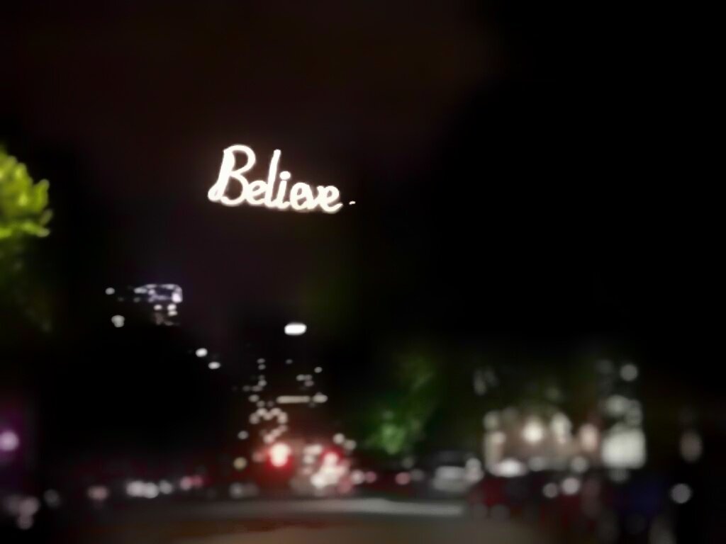 Federal Hill area in Baltimore at night. Pictured is a tree lined side street leading to a view of a tall dark commercial building in the distance. At the end of the street, above the trees and well in front of the commercial building, is a big, well illuminated sign that reads "Believe".

with a few cars along the lower third of the photo.
