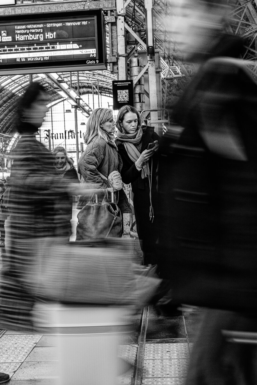 Two women look at the train app while people quickly walk past them.