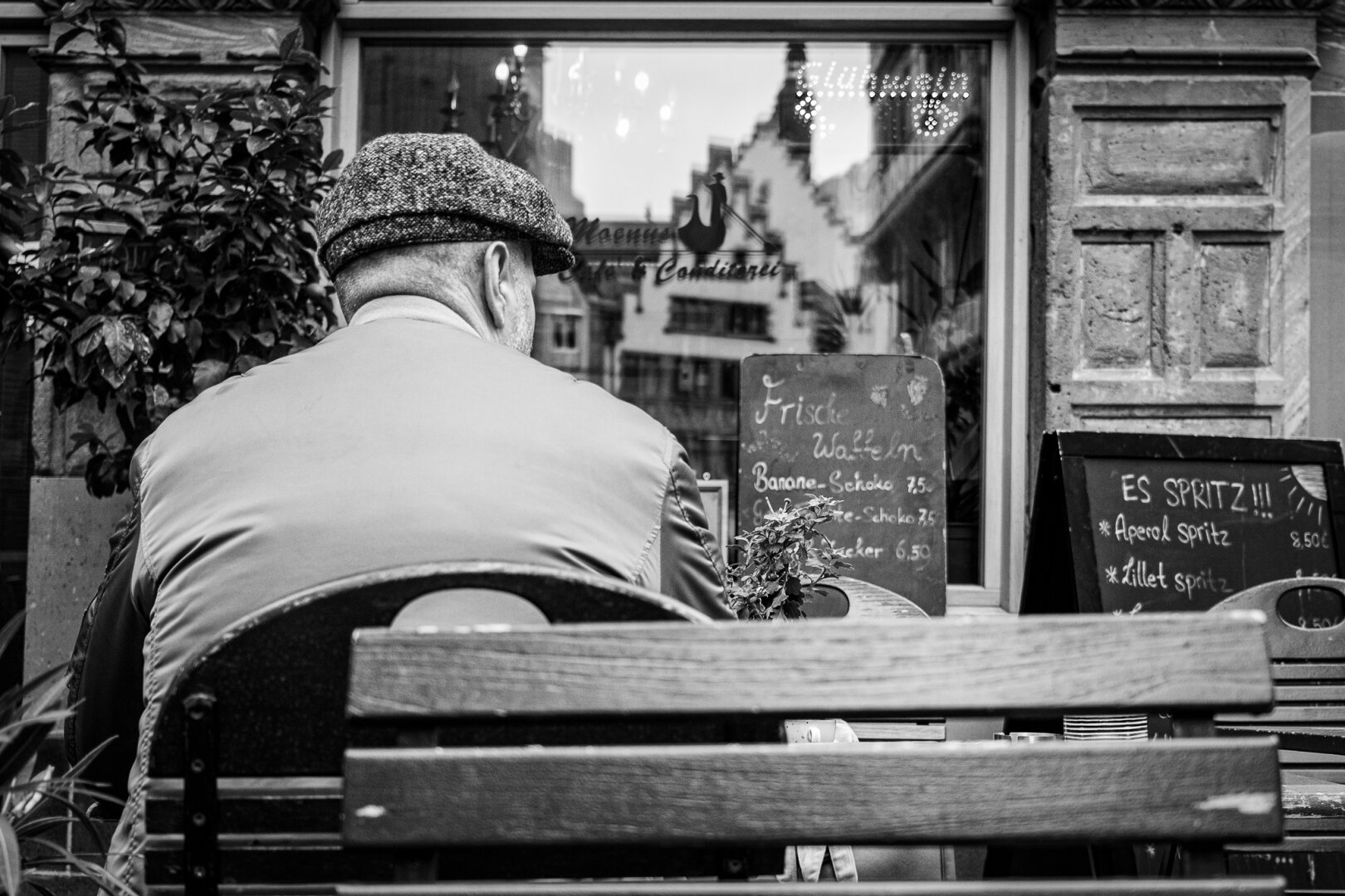 An elderly man sits with his back to the photographer in the outdoor seating area of ​​a cafe. The New Old Town is reflected in a shop window.