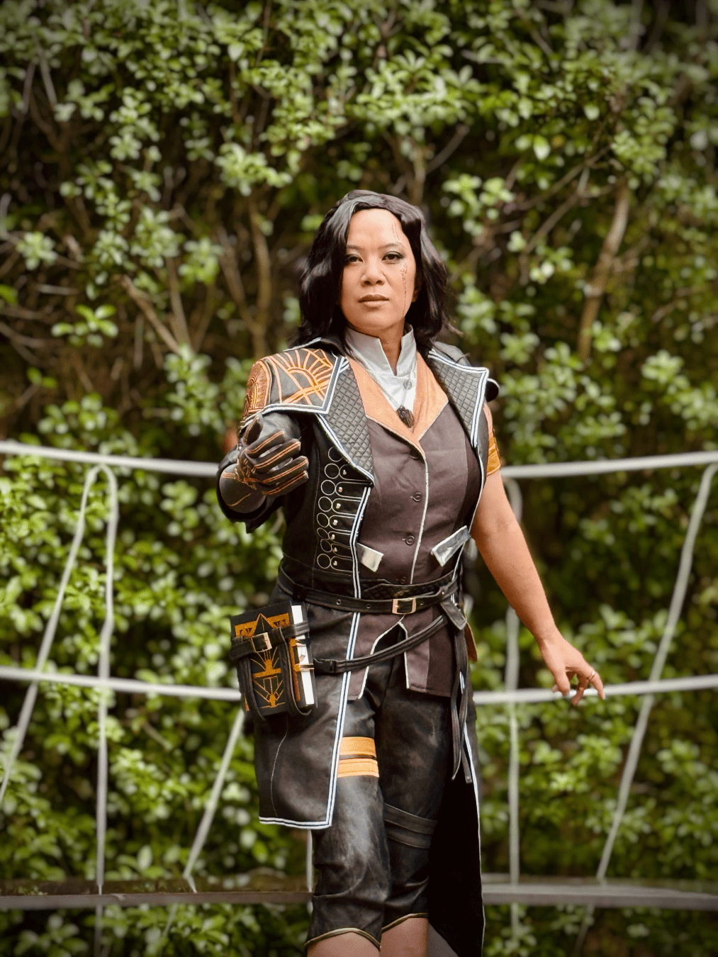 Cosplayer dressed as Lune from the game Clair Obscur: Expedition 33 in front of a wall of green leaves.