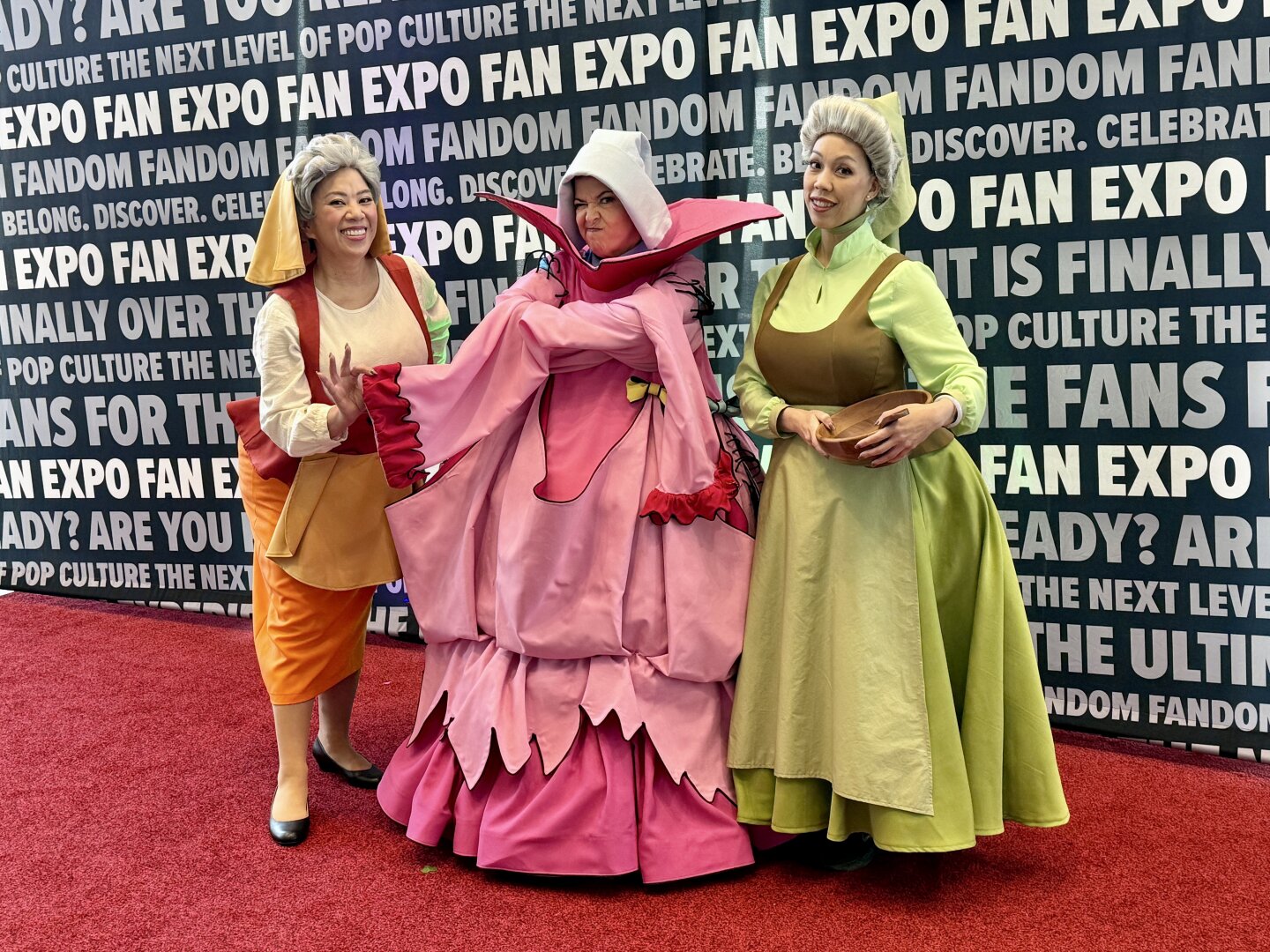 Three cosplayers dressed as Flora, Merryweather, and Fauna in their peasant costumes from the animated Disney movie Sleeping Beauty