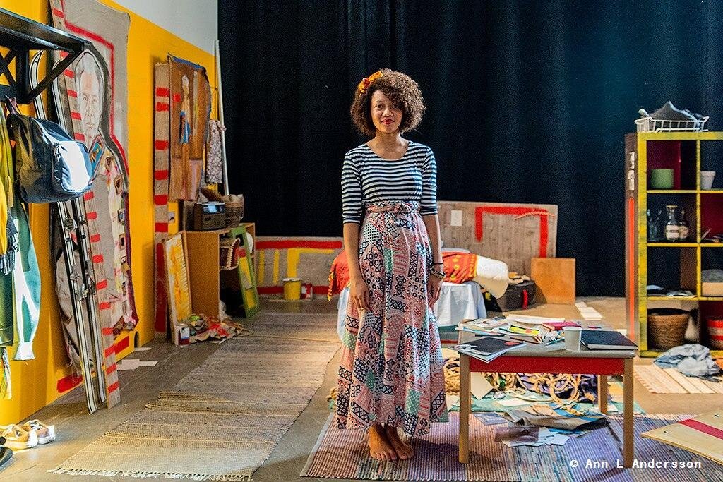 I'm standing in the middle of my installation work, a painting of my father to the right side with skis, a bed and cupboard behind me. On the right side of the bed is a painting of me as a child with a broken arm. On my left is a low table with notebooks and colouring pencils. I stand on a carpet with pieces of paper and photographs spread out on it.