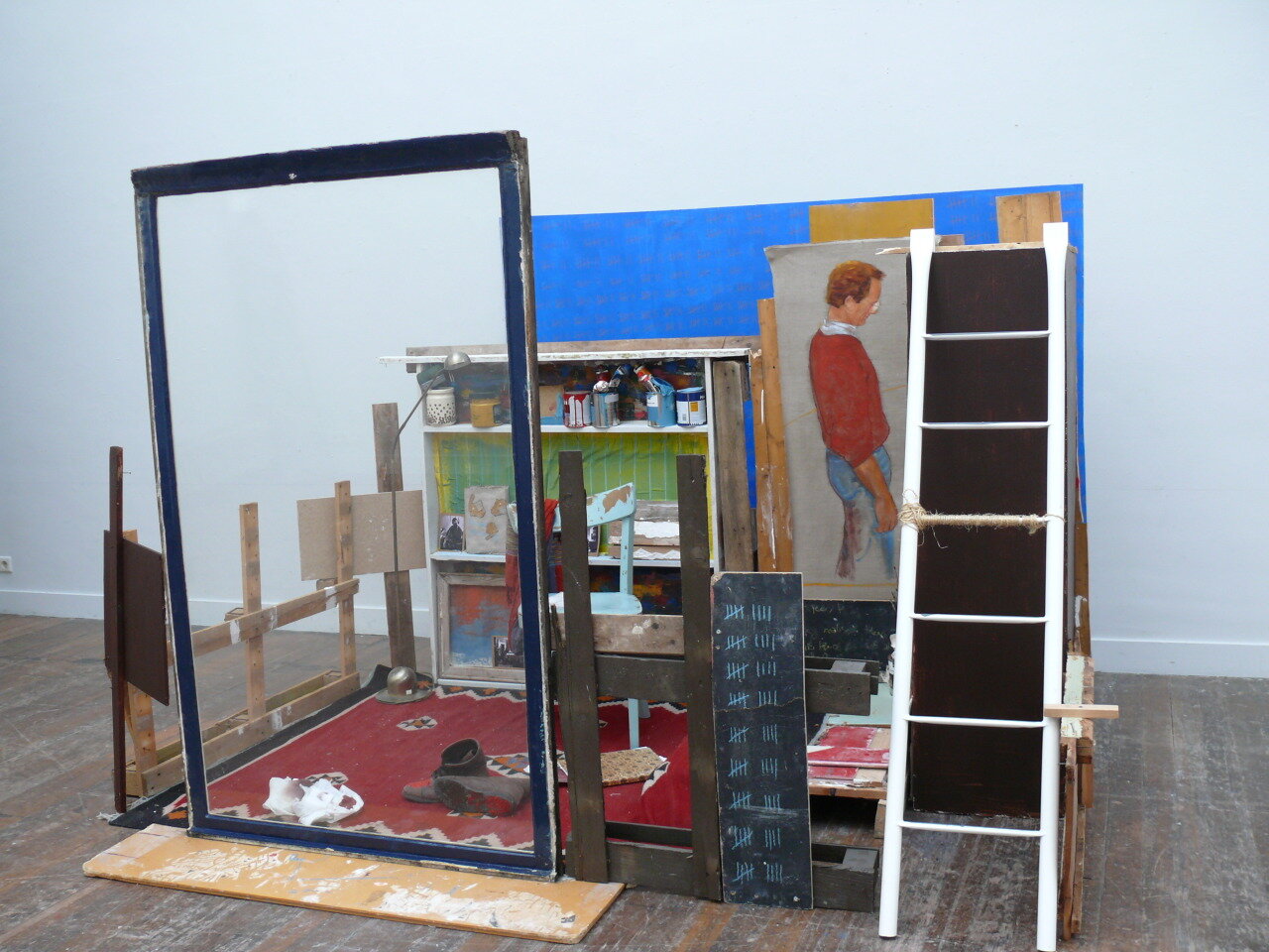 My installation for the master class at KABK, a small installation work with a glass window looking into the installation. A carpet on the floor, a bed staircase. A chair and my painting shoes on the carpet. A painting of my father in a red jumper and blue jeans.