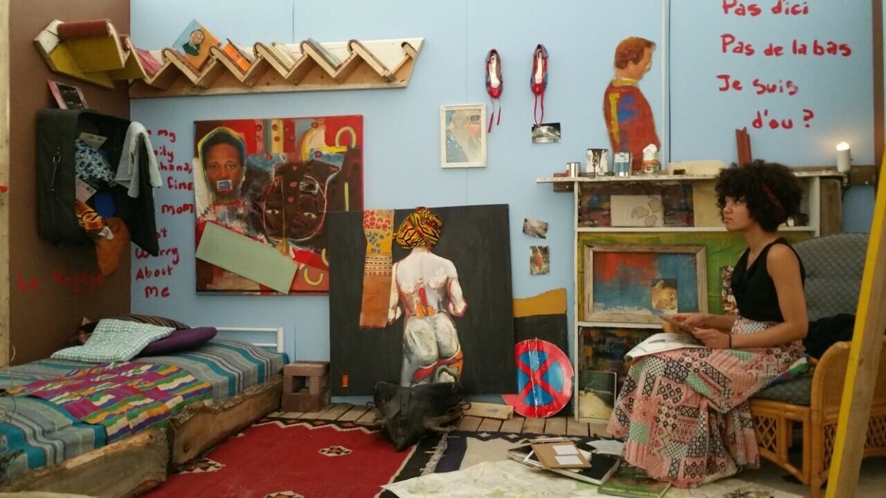 An Installation with me sitting on a chair. The words 'Pas d'ici, pas de la bas. Je suis d'ou?' written in red on the blue painted wall. There is a book shelf with a painting of my little brother on it. A painting of my father facing the red text. A painting of my mother with her mouth covered and below her is a painting of a woman's naked back with kente motif cloth wrapped on her head. 

A bed also has kente cloth on it and the suitcase of my ex is now hanging on the wall. a staircase makes for a bookshelf stuck up on the wall above the bed.