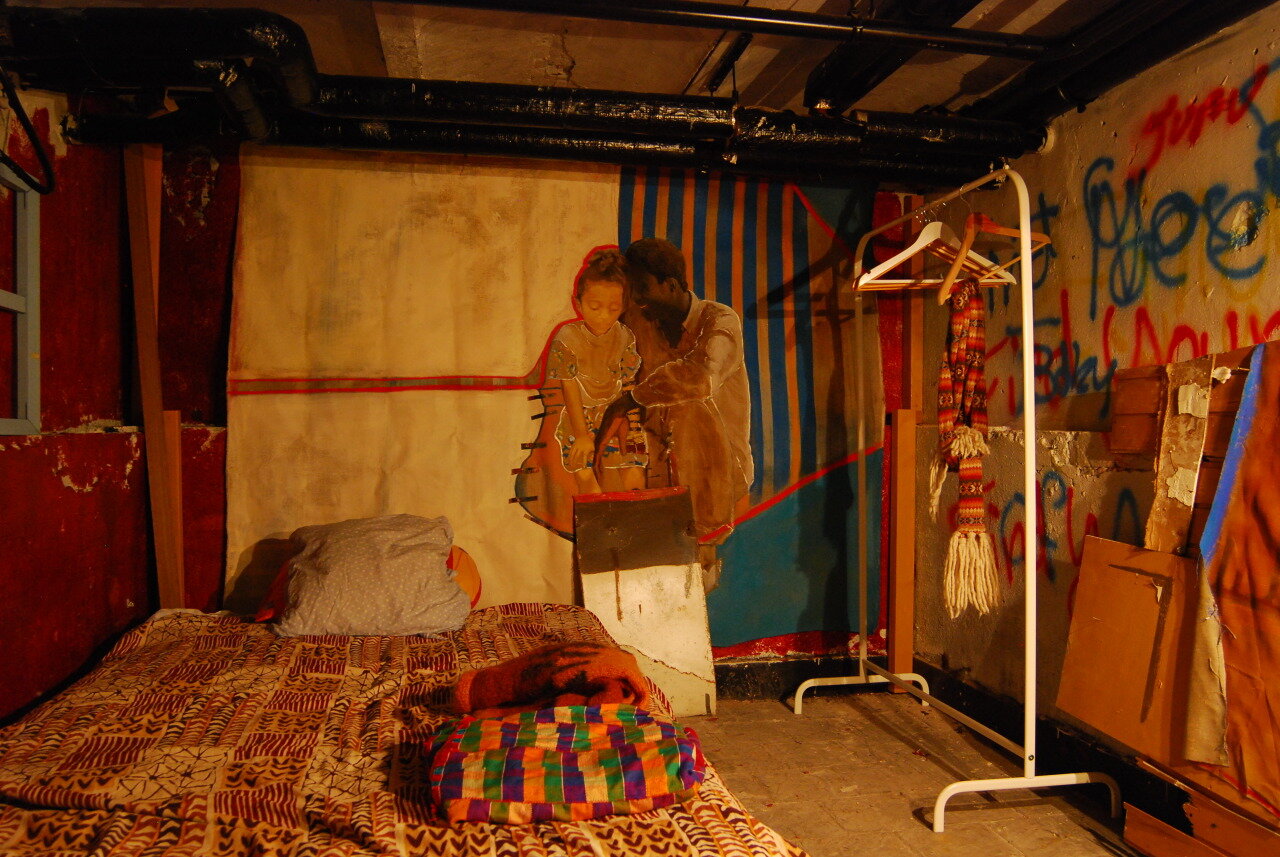 My graduation work at KABK sets for a bedroom with a bed with kente cloth covering it. A large painting of myself as a child sitting on the lap of a family friend with a light background. A cloth hanger stand next to it.