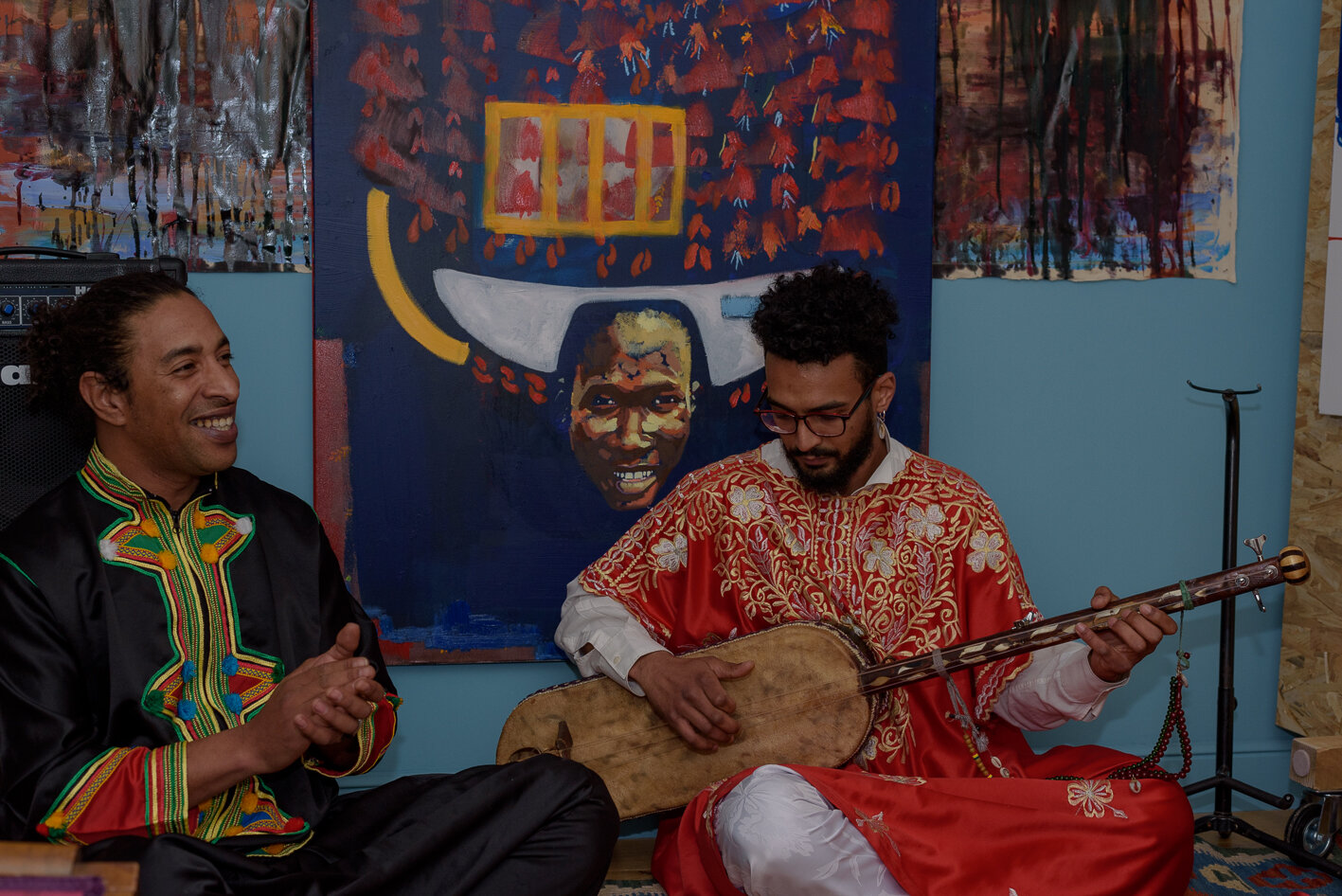 Two of the three Gnawa musicians. One is in red holding and strumming guembri instrument and the other in black is clapping