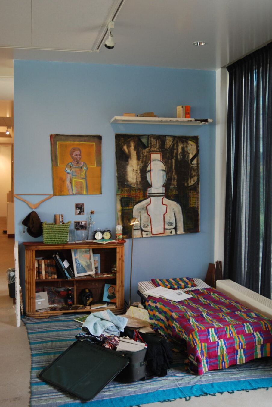 An installation work with a bed with Ghanaian kente cloth. next to the bed is the suitcase of my ex boyfriend. on the light blue wall are two paintings, one of my grandmother with the text 'forgive me' written on her clothes and next to her is a painting of a woman figure. A bedside table holds two clocks with the same time on them.