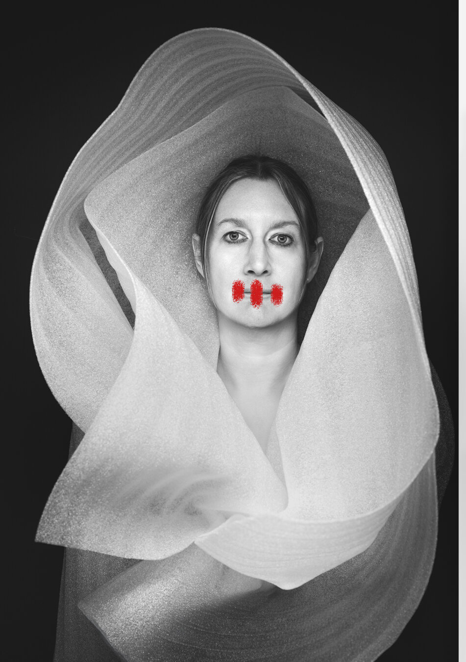 Black and white headshot of a woman who is dressed in white oversized hood made of protective packaging material. She is not looking directly into camera, there are three vertical red marks across her lips as if her mouth is sewed so she can’t talk