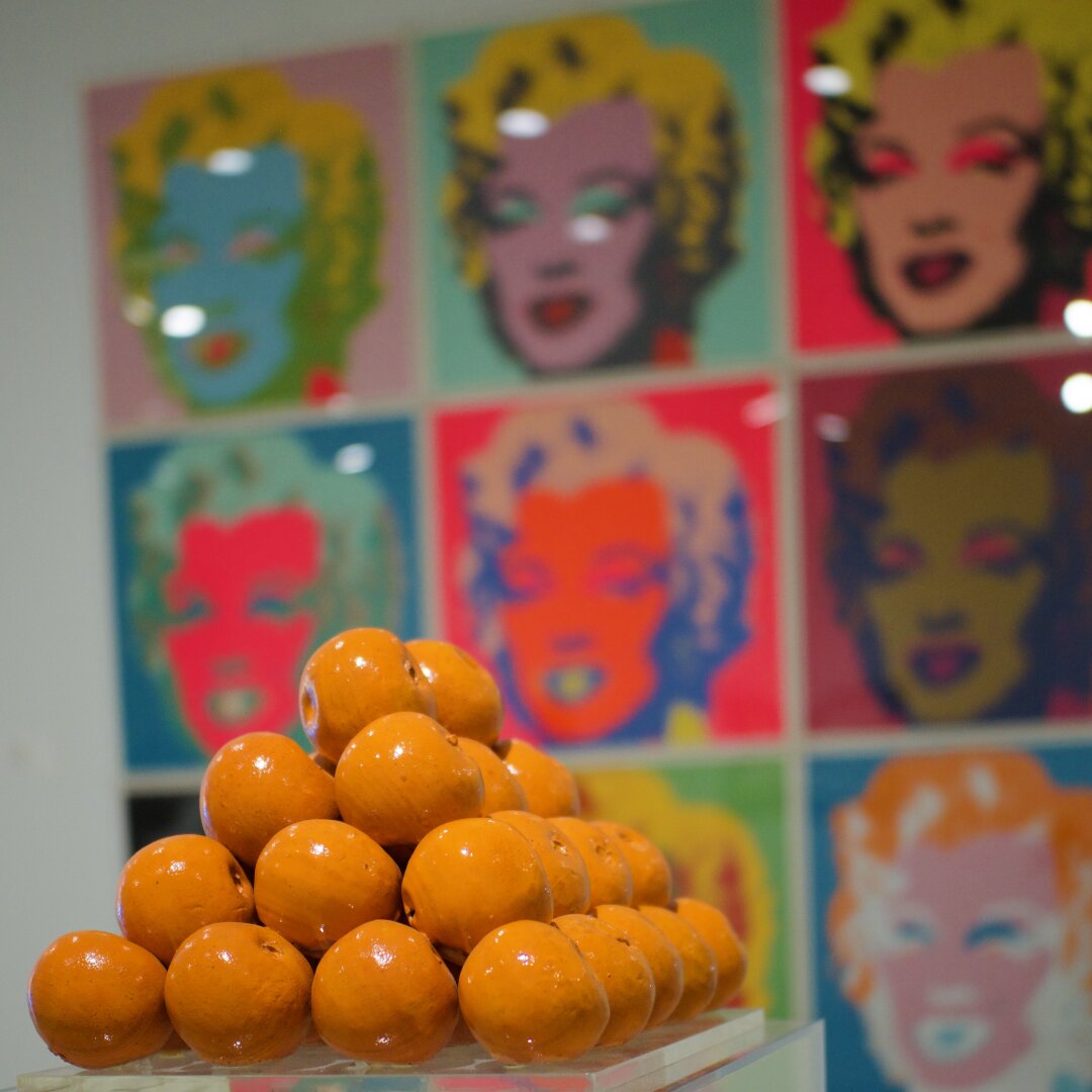 A bunch of shiny stylized orange apples in the foreground. Andy Warhol's "Marylin Monroe" on the wall in the background.