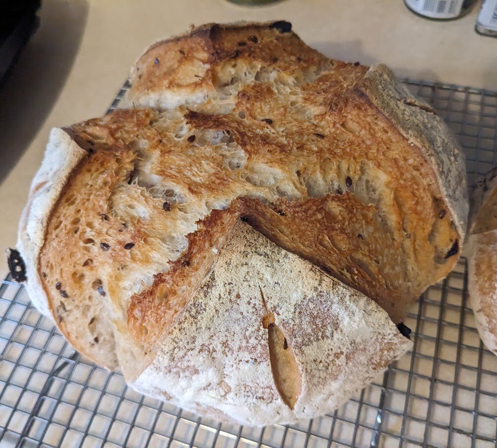 A large sourdough bread boule, slashed with a simple X-pattern. Small dark pieces of black garlic are visible on the surface.