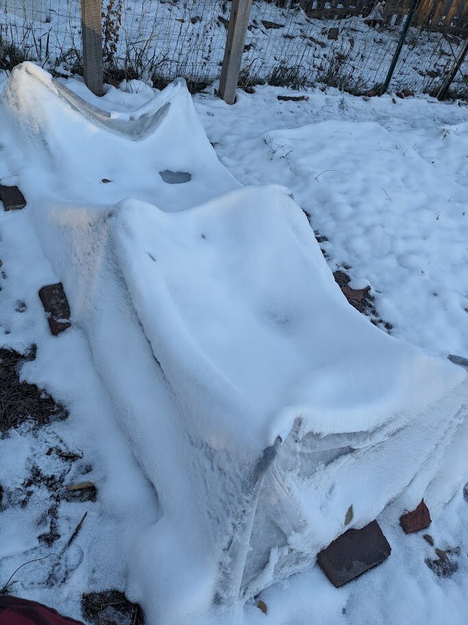 A small raised bed covered in hose-hoops and frost cloth is buried in snow. The frost fabric is weighed down and sagging in the middle, which isn't ideal.