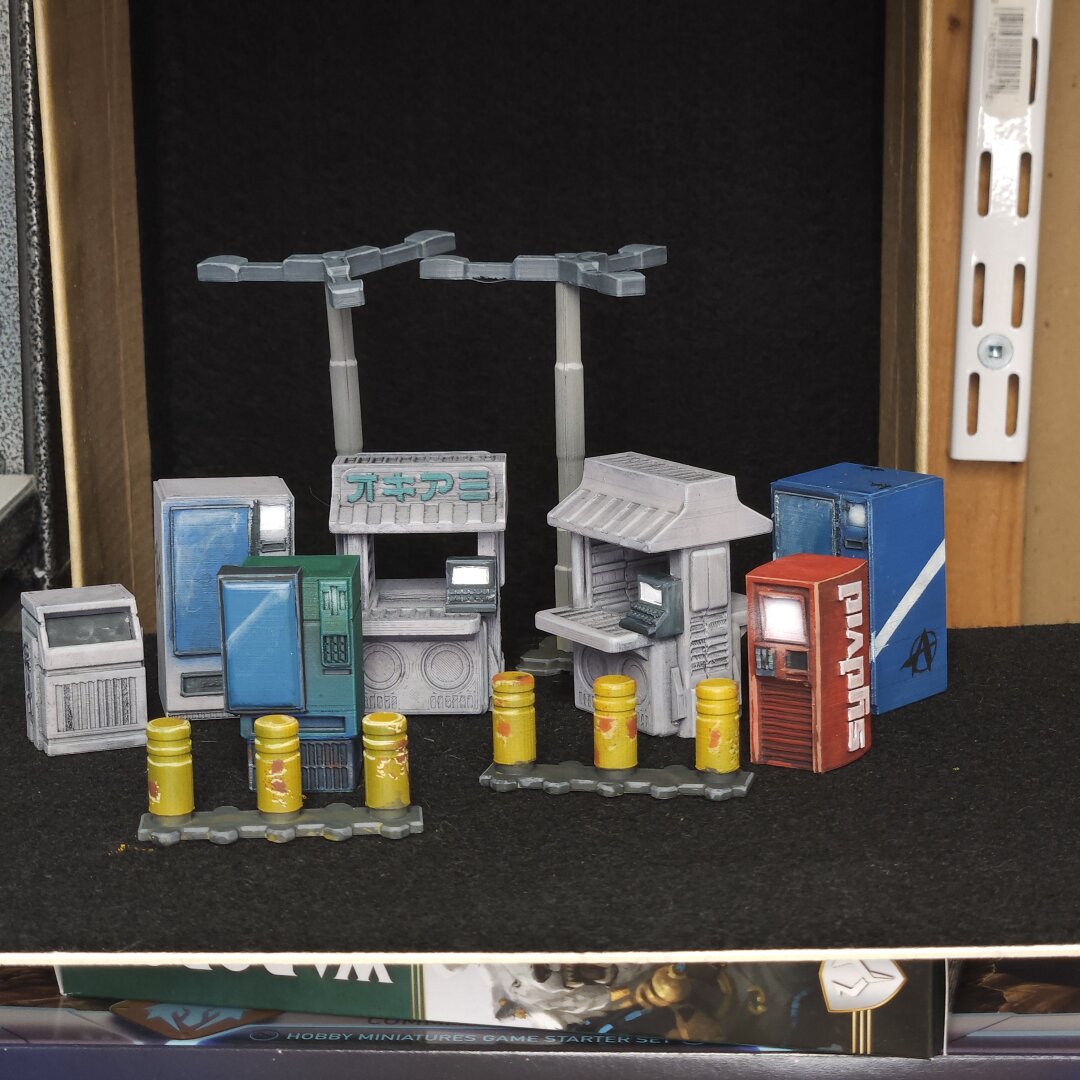 A photograph of some miniature wargame terrain. The terrain is 3d printed vending machines, lamp posts and traffic bollards. The terrain is from Corvus Games Terrain and it was printed and painted by Tyler Provick.