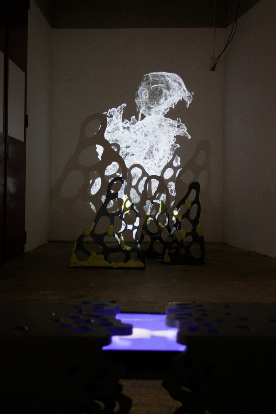 salle d'exposition avec une projection sur structure de bois en fondn au premier plan un banc noir à l'intérieur du quel se trouve un écran montrant une image bleue