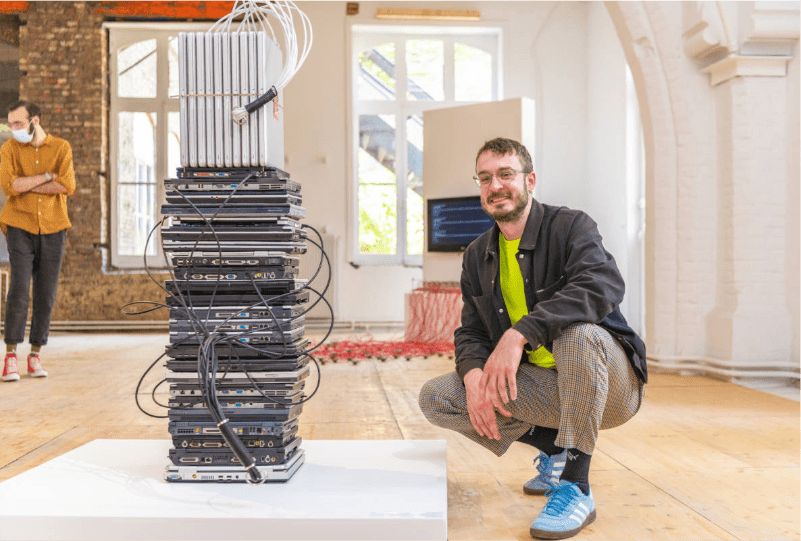 photographie de l'artiste marcel schwittlick à côté de sa scultpure, composée d'une pile d'ordinateurs gris et noirs