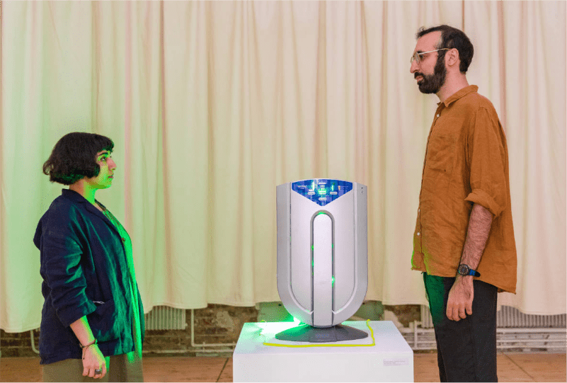 un stérilisateur d'air est posé sur un socle entourée d'une lumière verte, de chaque côté les deux artistes : Maryam Katan et Shaahin Peymani