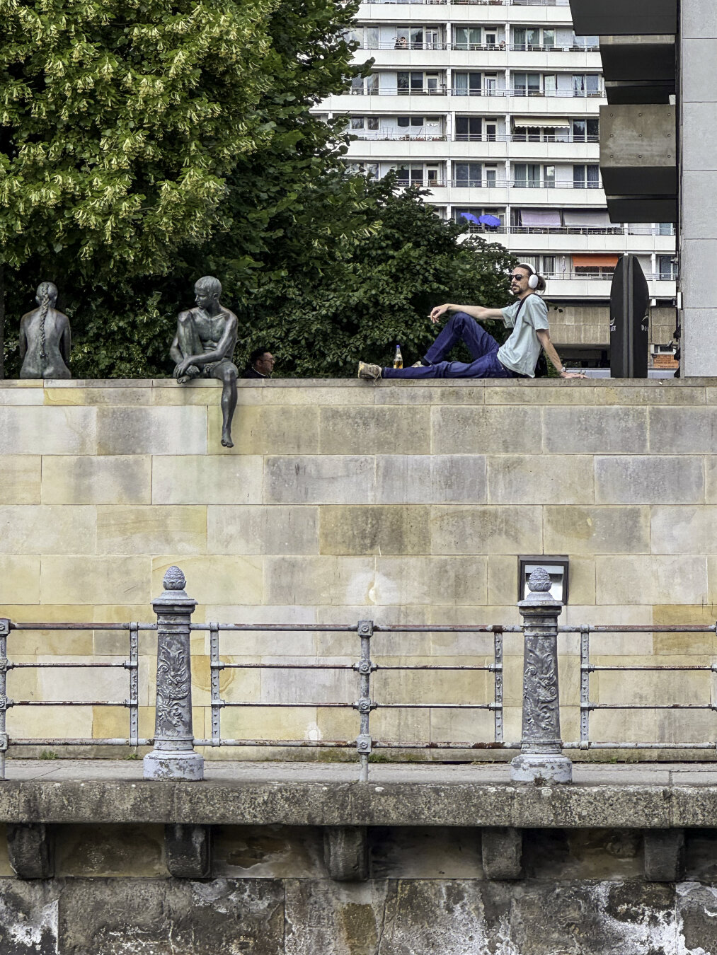 A man sitting on a wall wearing headphones next to two bronze sculptures of human figures.