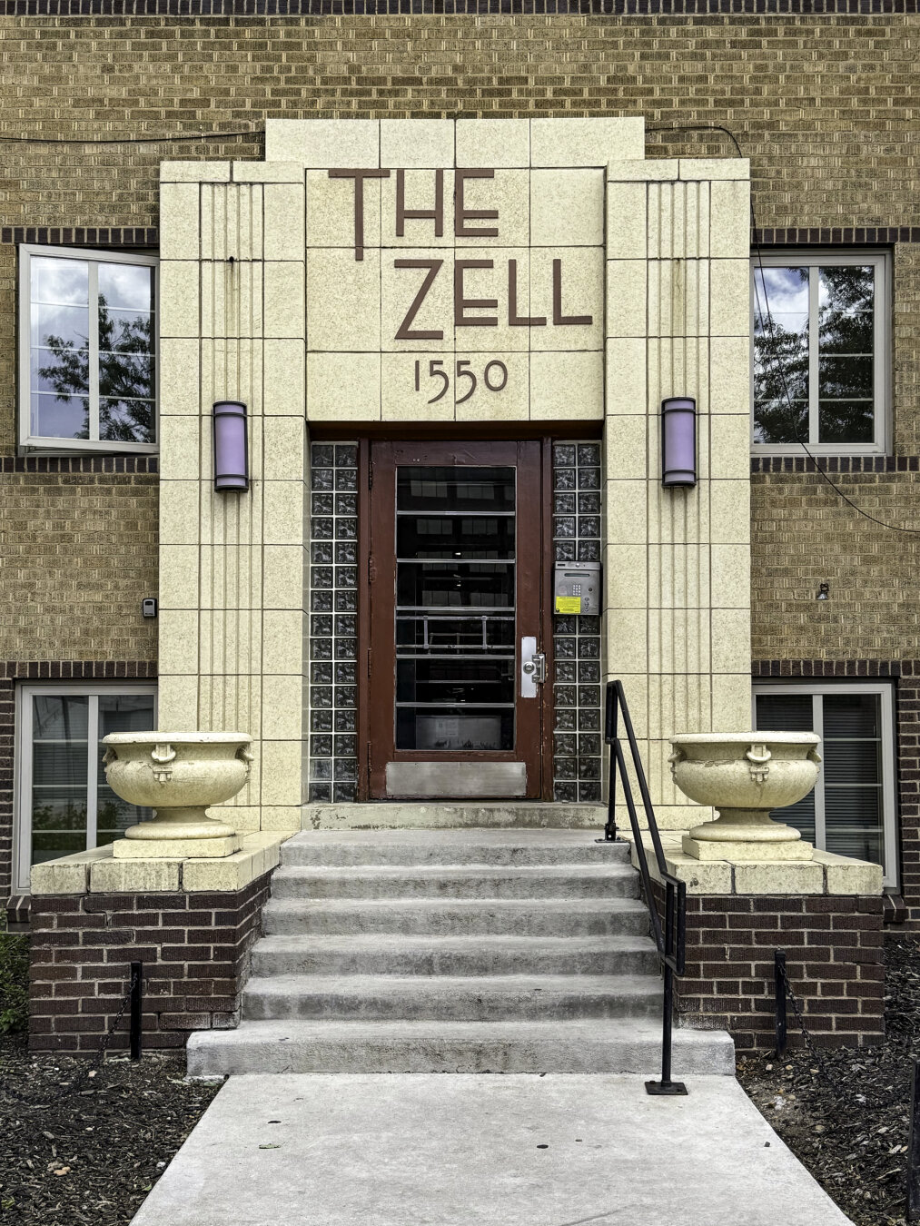 A building entry consisting of a red door surrounded by a pale stone geometric surround in a late Art Deco style with fluted vertical bands. Two classical style urns sit on short walls on either side of the steps leading to the door. In the center of the surround above the door in large Art Deco typeface reads the building name, THE ZELL.