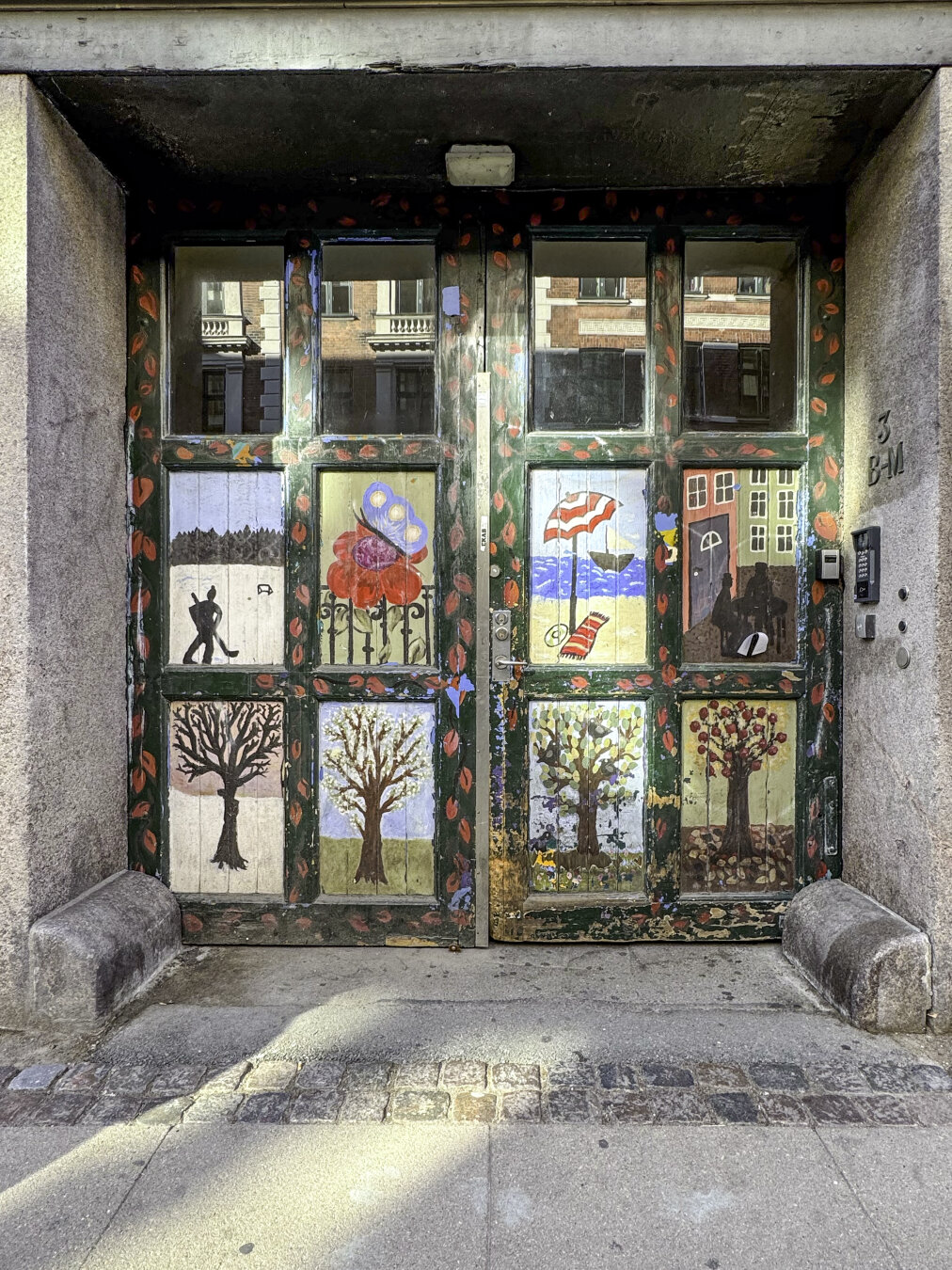 A double wooden door with multiple square panels, each painted with an artistic image, deeply recessed within a masonry portal.