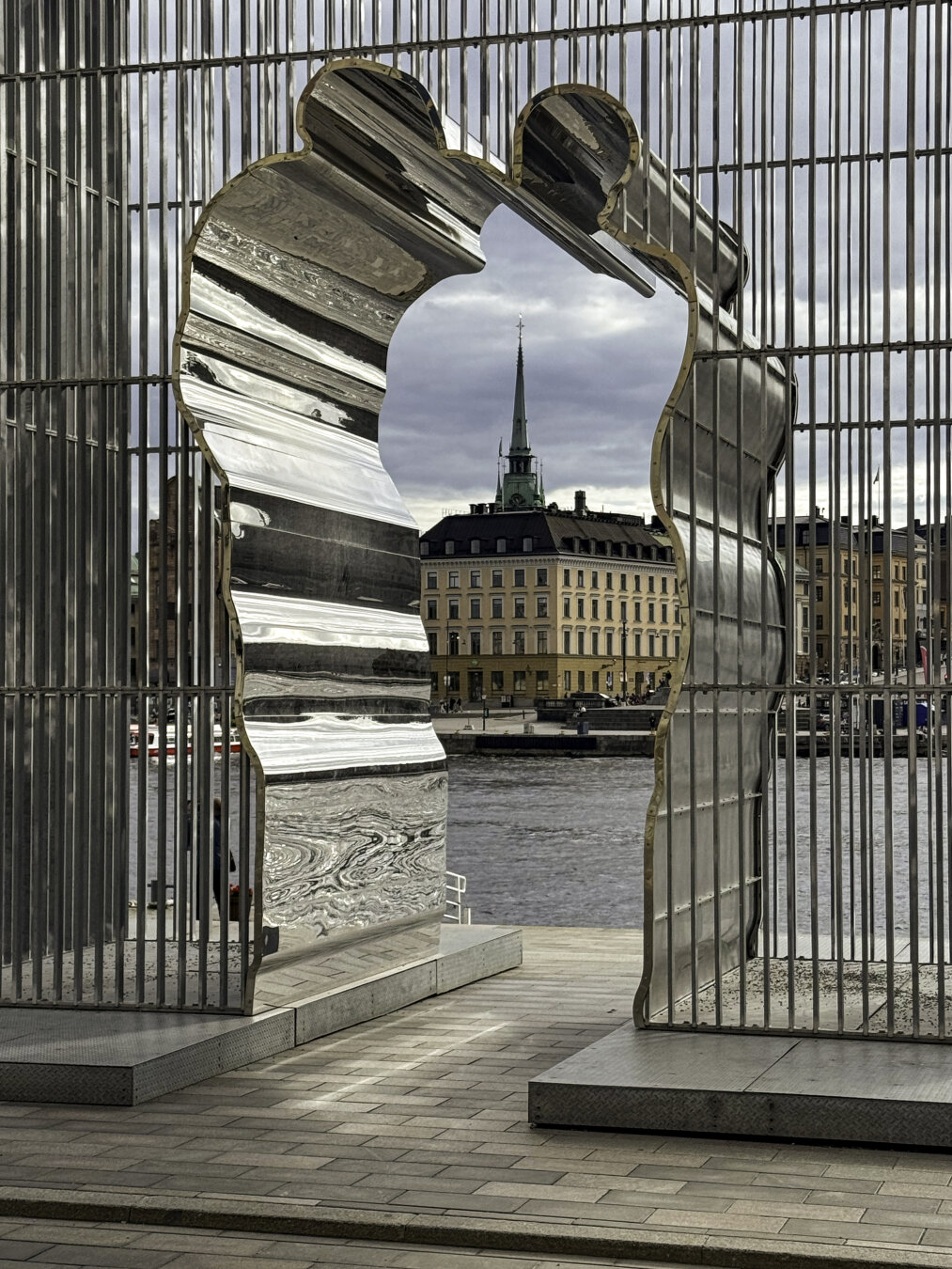 Stainless steel arch in an irregular shape with the historic buildings beyond and water in the foreground.
