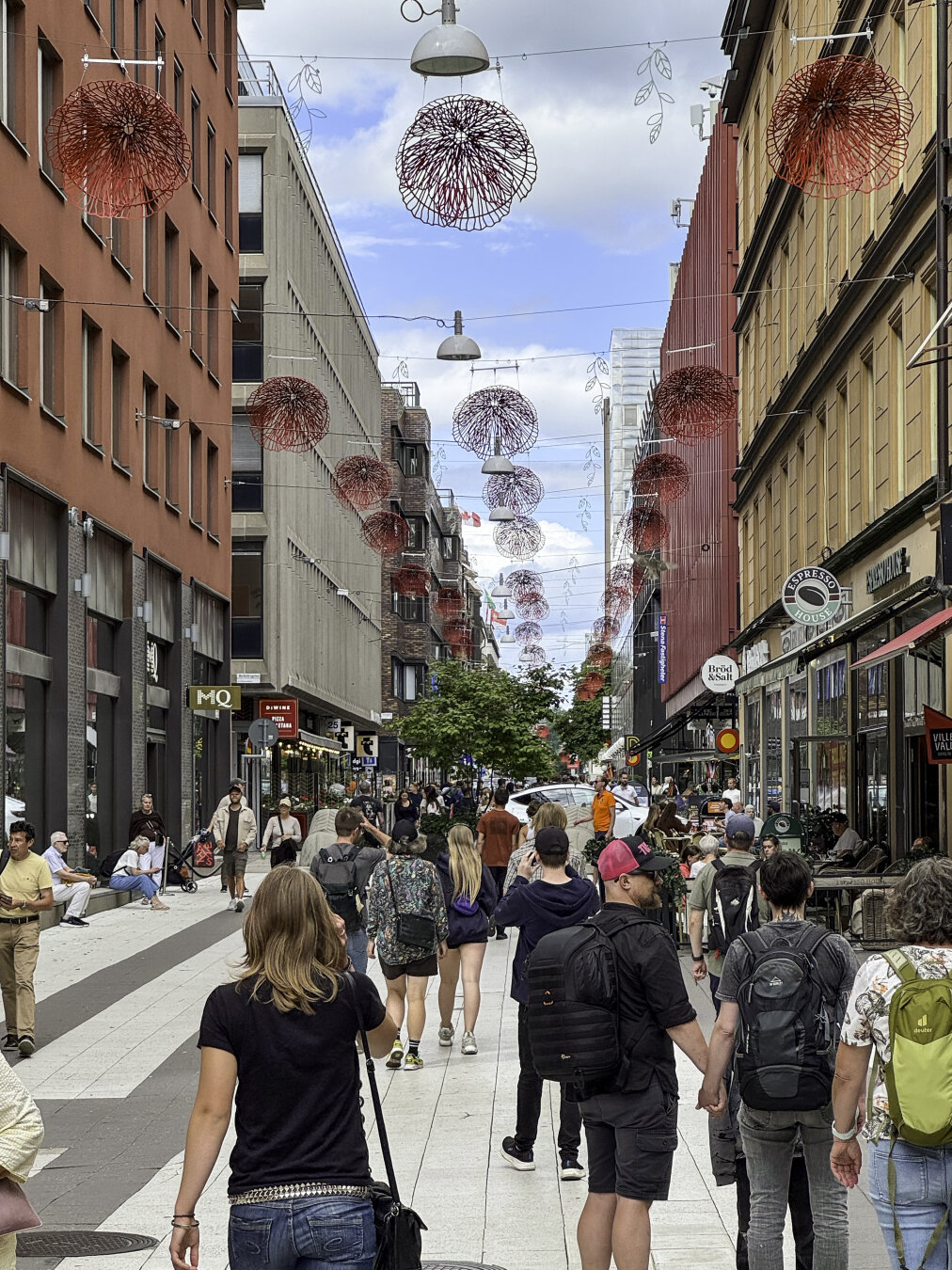 Dozens of pedestrians walk along a pedestrian-only street with storefronts lining both sides and red artistic flowers hanging from wires above the street.