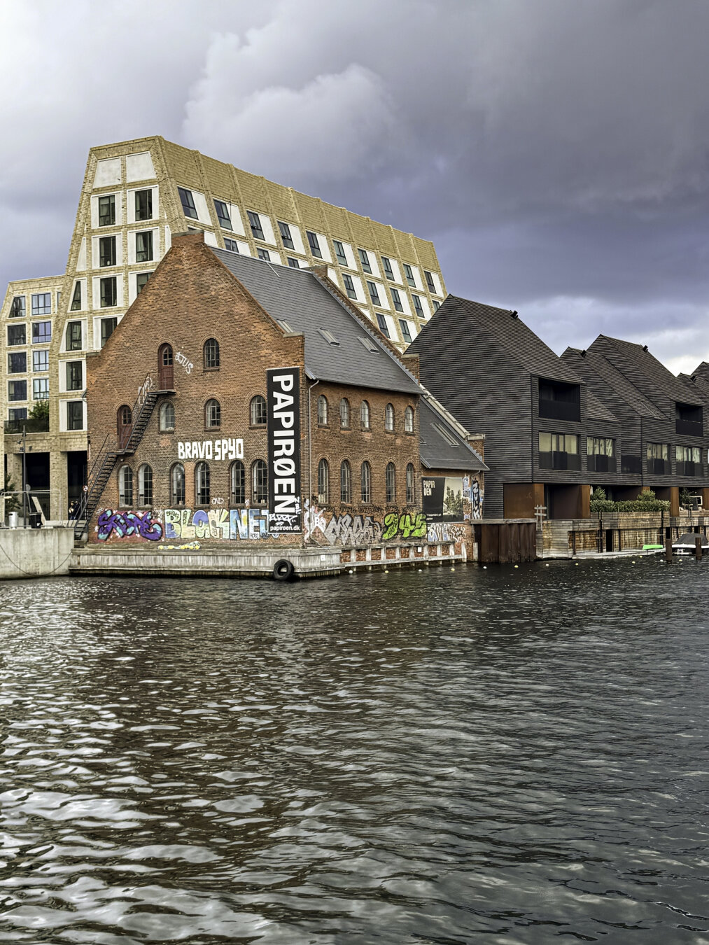 A historic brick building with a peaked room next to modern buildings with designs that take on the same form.