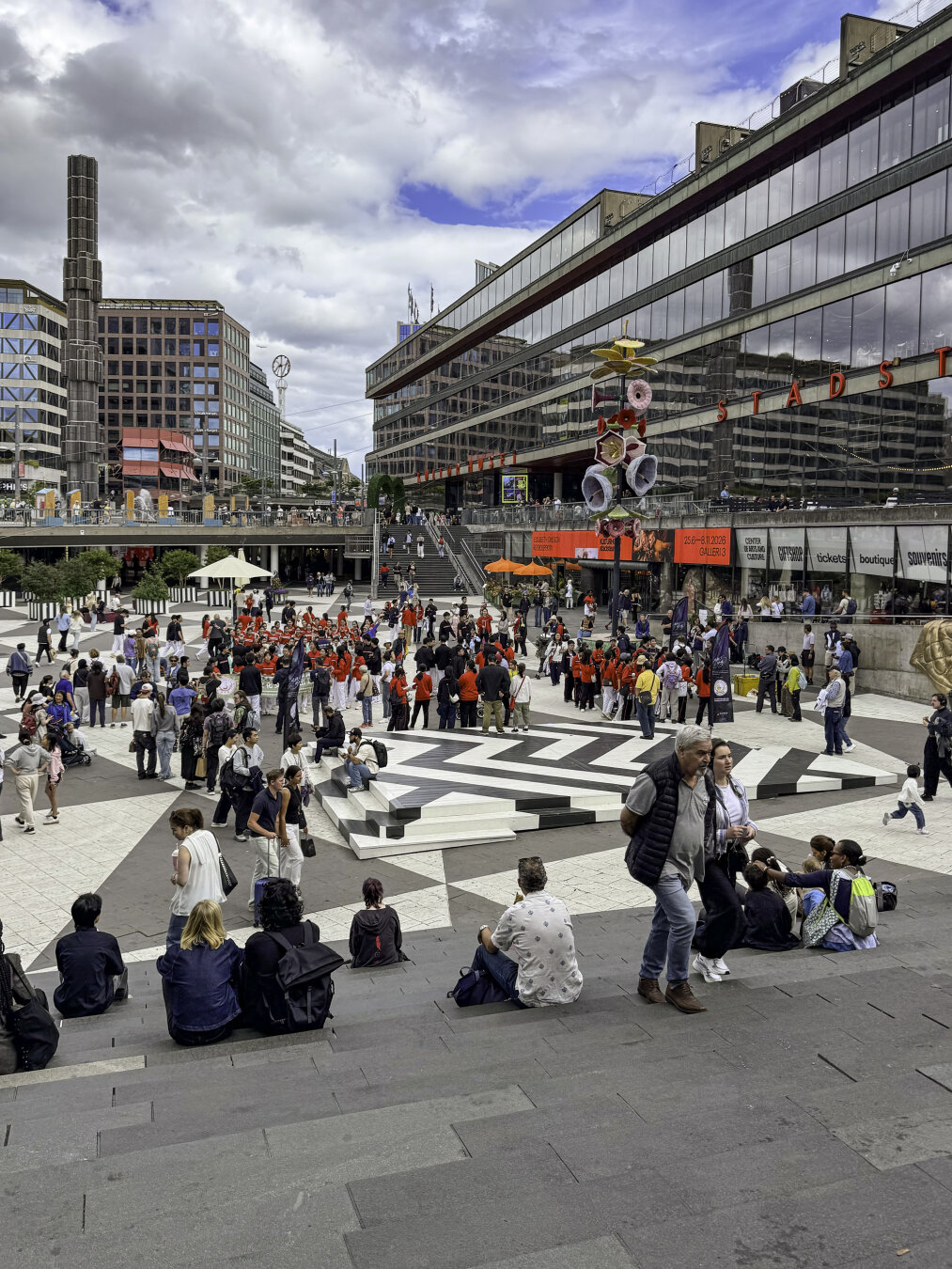 Dozens of people are gathered and hanging out in a large modern plaza surrounded by modern buildings. The plaza is sunken below street level with stairs leading up to the street.