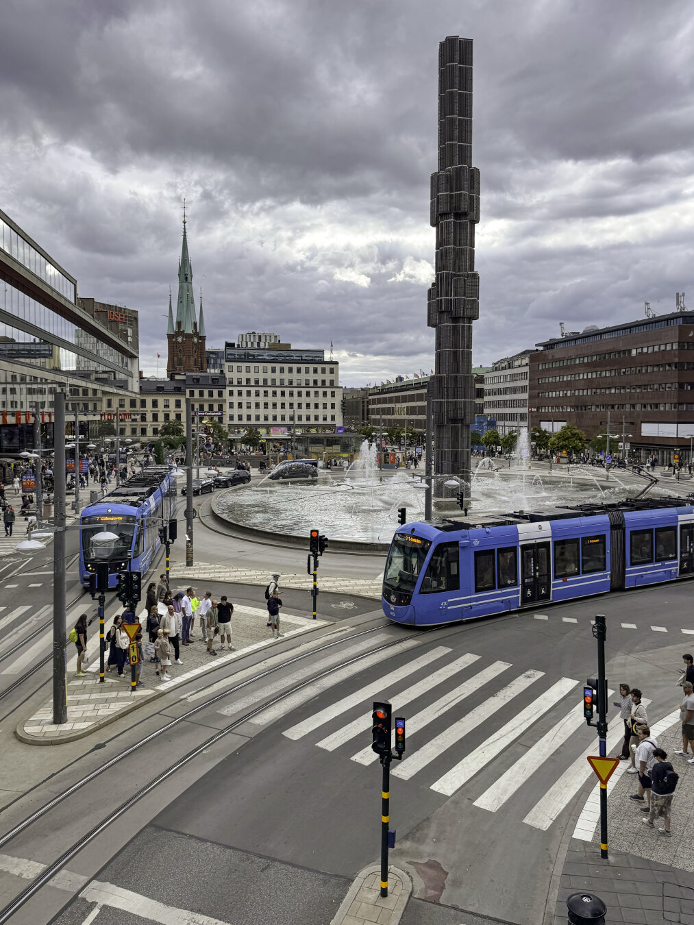 Large public plaza with an artistic obelisk in the center of a large fountain, and two blue public transit trams moving through the space on the public streets along the edge.