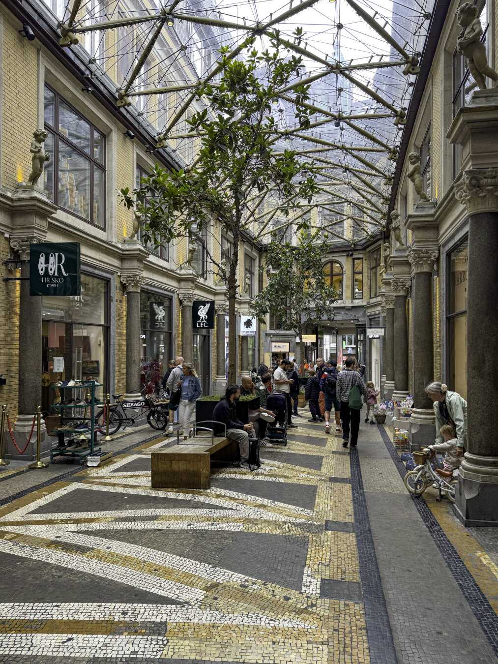 Pedestrians hang out in a glass-enclosed street lined with shops.